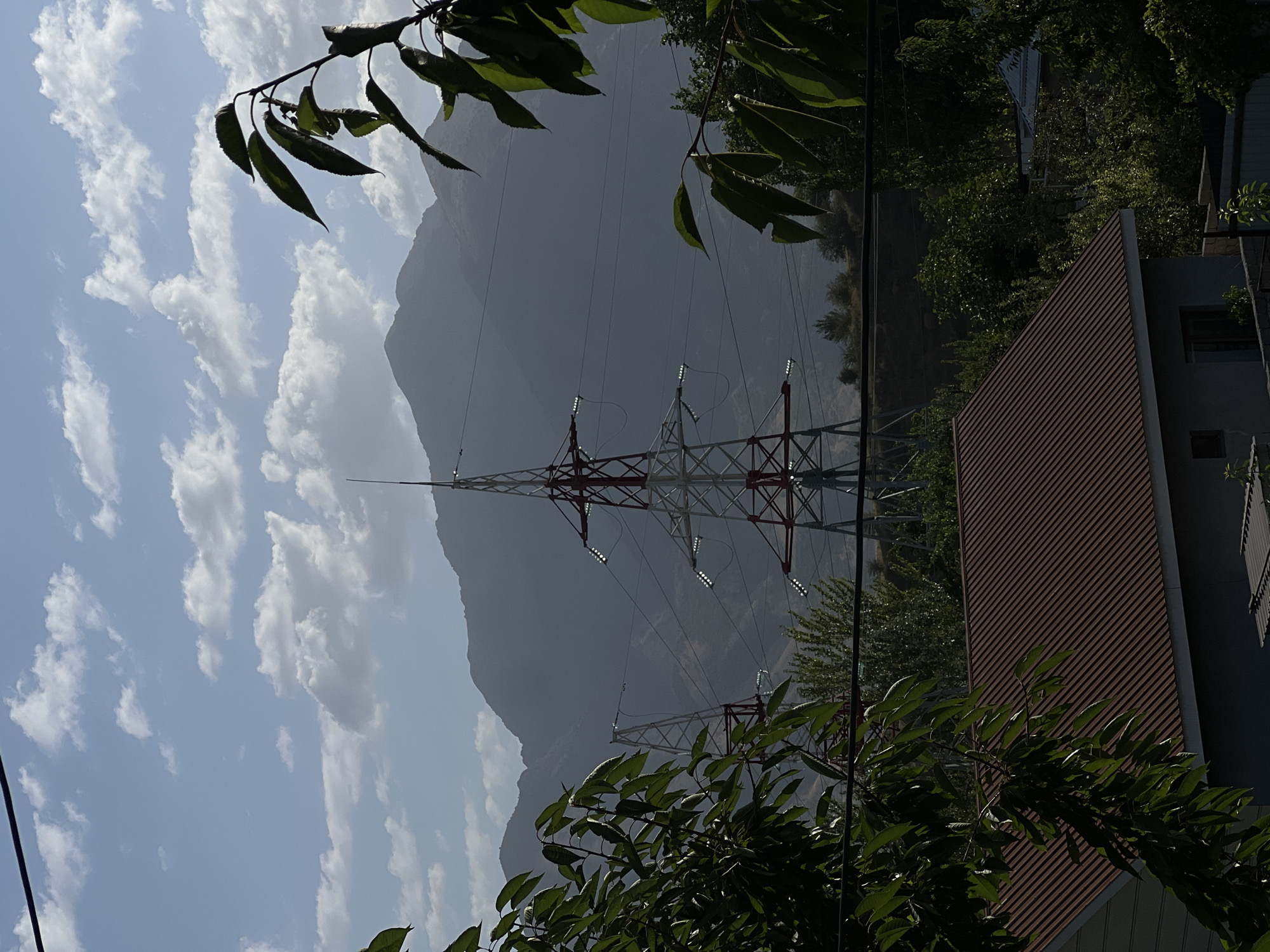 A medium shot captures a residential area in Yusuphana, Uzbekistan, framed by foliage in the foreground and a mountainous landscape in the background. In the mid-ground, two towering electrical transmission towers, painted with red and white accents, stand prominently. These towers are connected by thick power lines, some of which are equipped with what appear to be lights. Beyond the towers, a range of hazy, muted mountains stretches across the horizon under a sky dotted with soft, white clouds. In the lower portion of the image, a house with a reddish-brown corrugated metal roof is partially visible, nestled amongst lush green trees and vegetation. The overall scene depicts a juxtaposition of rural and industrial elements within a natural setting.