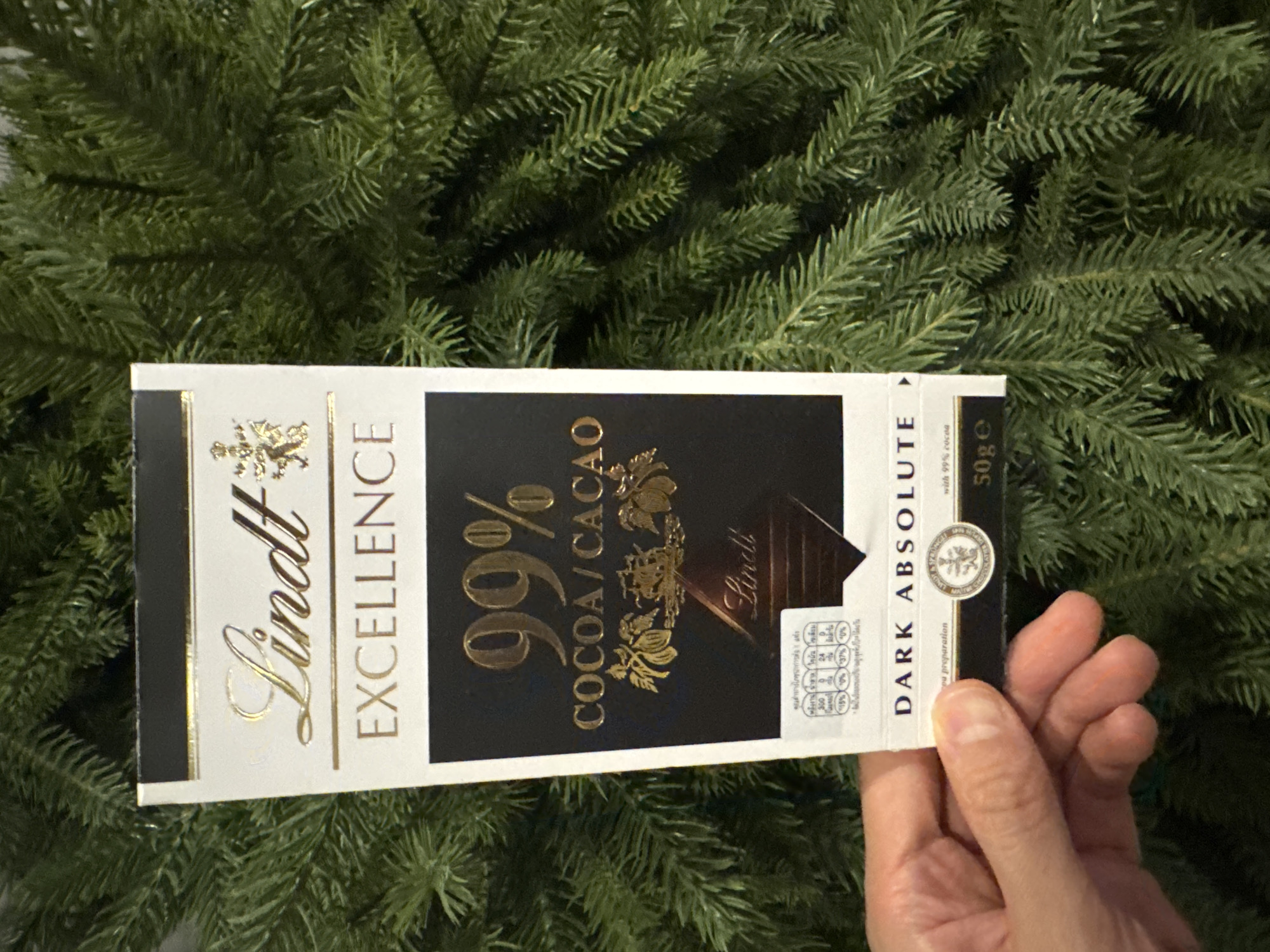 A close-up image captures a hand holding a Lindt Excellence 99% Cocoa/Cacao Dark Absolute chocolate bar vertically. The hand, with visible fingernails, is positioned on the lower right side of the chocolate bar. The chocolate bar's packaging is predominantly black and white with elegant gold lettering. Prominent text includes "Lindt Excellence" on the left, "99% COCOA/CACAO" in the center, and "DARK ABSOLUTE" on the right. Further details like "with 99% cocoa" and "50g e" are also visible. On the right side of the bar, there's nutritional information, including the Thai phrase "คุณค่าทางโภชนาการต่อ" (Nutritional value per) followed by figures such as 300 and 24, and various percentage daily values. The background consists of a dense, out-of-focus array of green, needle-like branches, characteristic of an artificial Christmas tree or pine garland. The setting appears to be indoors. The presence of Thai text on the packaging indicates the location context is Bangkok, Thailand.