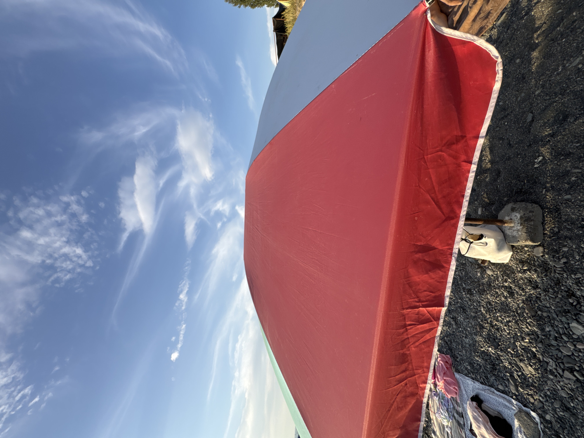 A low-angle, close-up shot captures a large, multi-colored beach umbrella dominating the foreground. The umbrella canopy is divided into sections of red, light blue, and a sliver of green at the top left. The fabric of the umbrella is wrinkled, indicating it is deployed. The umbrella is supported by a metal pole anchored in a concrete block on a gravelly ground. Scattered on the ground beneath the umbrella are various items, including folded clothes, a white bag, and a piece of fabric. The background consists of a bright blue sky with wispy white clouds and a hint of distant trees and structures. The scene suggests an outdoor setting, likely a beach or park, under a clear sky. The location is specified as Zharbulak, Kazakhstan.