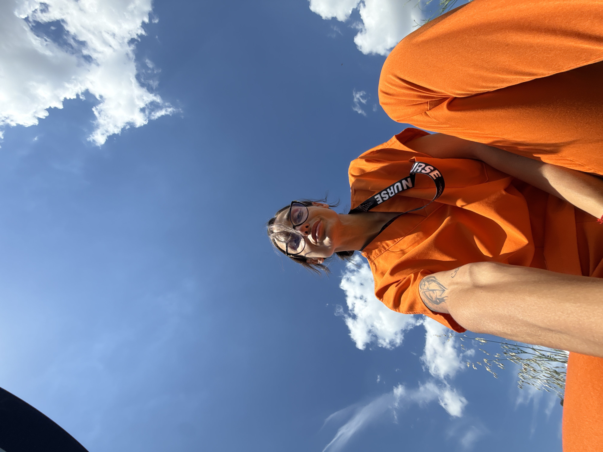 A low-angle, wide shot captures a person wearing bright orange scrubs, possibly a nurse, against a clear blue sky dotted with white clouds. The person is looking up at the camera with a slight smile and wearing glasses. A lanyard with the word "NURSE" is visible around their neck, and a tattoo is present on their left arm. The lower half of the frame shows their legs in matching orange scrub pants. The scene appears to be outdoors, with some dry grass visible in the lower left corner. There is no specific information in the image that would indicate the location in the city of Pleven, Bulgaria.