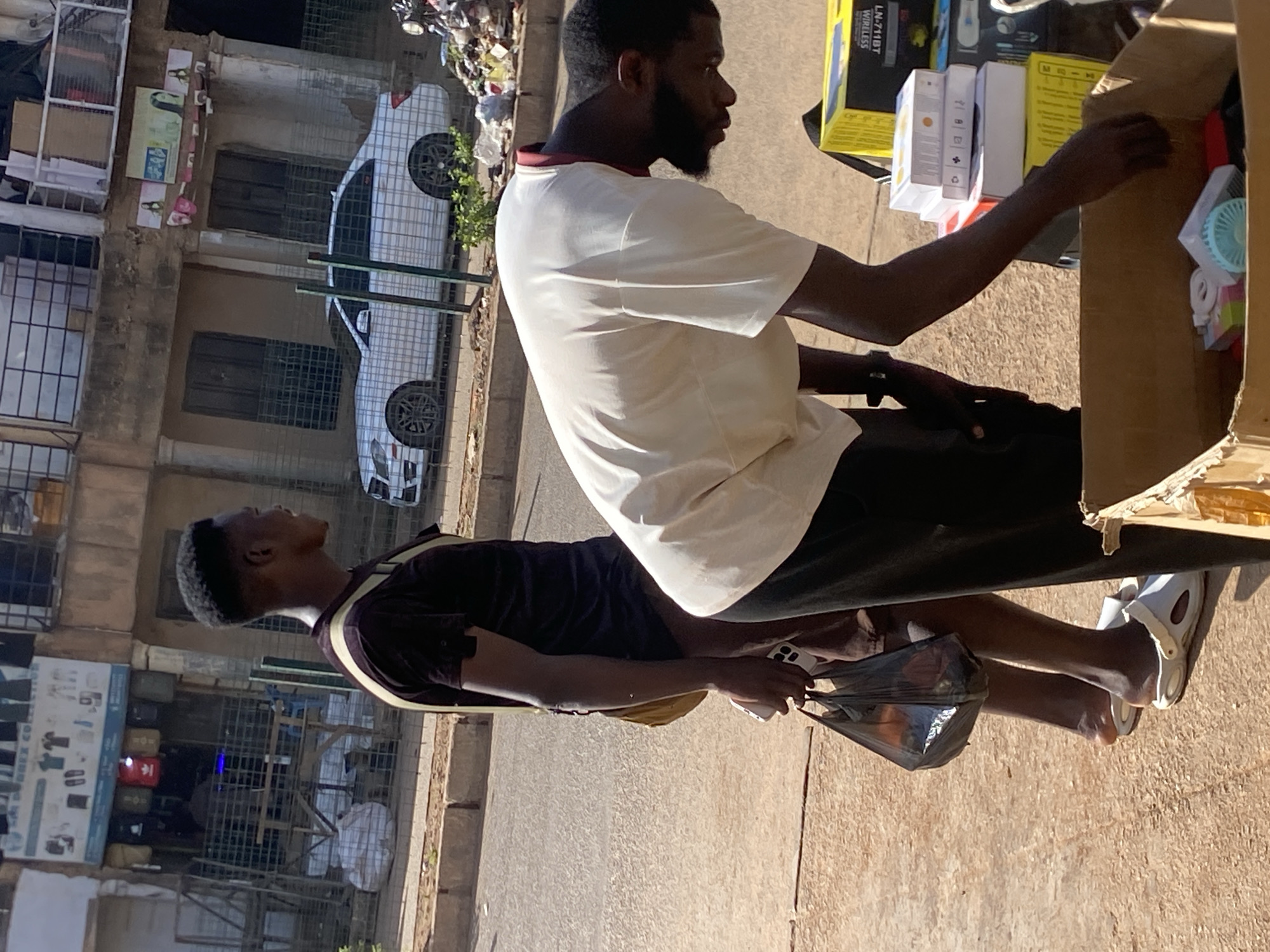 In Bukuru, Nigeria, two men are seen on a street. One man, wearing a dark maroon shirt and dark pants, walks carrying a black plastic bag in his left hand. He is also wearing a light-colored shoulder bag and white slides. The second man, wearing a light-colored t-shirt and dark pants, is bent over a cardboard box filled with various items, including small electronic devices and boxes. In the background, a white sedan is parked on the street, and a building with shops and balconies is visible. The ground is paved with concrete and asphalt.