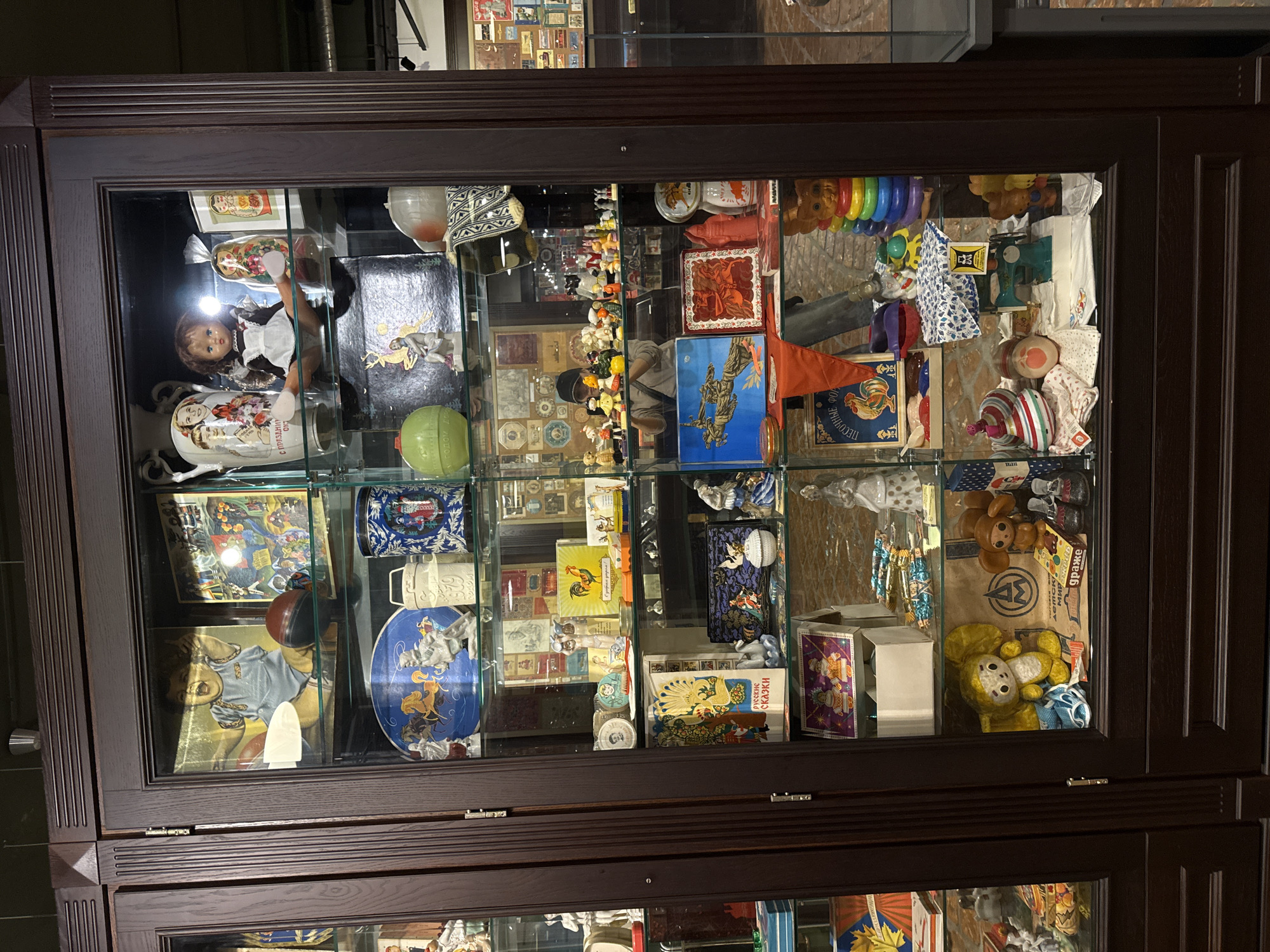 This image shows a glass-fronted cabinet filled with a collection of vintage toys and decorative items. The cabinet is dark wood and appears to be in a museum or exhibition setting.
The top shelf displays a large framed picture of a baby, a ceramic jug with images of children, a doll in a school uniform, and several nesting dolls.
The second shelf contains a decorative plate with horses and figures, white ceramic milk jugs, and a green ceramic object.
The third shelf is crowded with small ceramic figurines of people and animals, several small boxes, and a yellow box with a rooster on it.
The fourth shelf features a book with a peacock illustration, small decorative boxes, a white ceramic ballerina, and other small trinkets.
The fifth shelf holds a colorful matryoshka doll, a stack of colorful rings, and several stuffed animals.
The bottom shelf contains a large yellow stuffed Cheburashka toy, another stuffed toy resembling a monkey, and a box labeled "ДРАЖЕ" (dragees).
The background of the cabinet reveals a blurred image of the exhibition space, including other display cases and some pipes on the ceiling.
The text on some of the items is in Russian. The style of the toys and decorations suggests they are from the Soviet era.
The location is specified as Arkhangel'skoe, Russia, suggesting this is a display within the Arkhangelskoye Estate Museum.