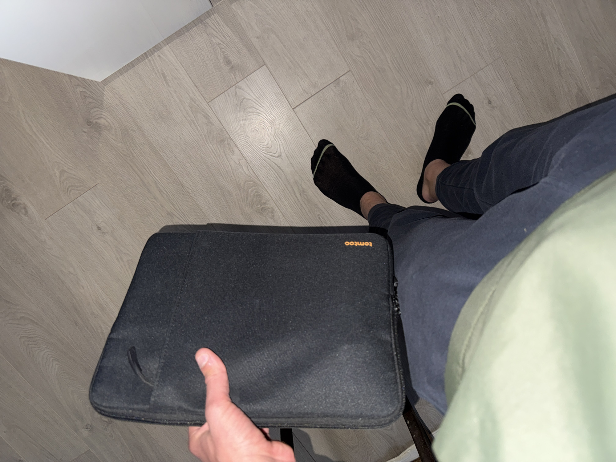 The image shows a person sitting on a chair in an indoor setting, possibly a home or office. The floor is covered with light-colored wood-like flooring. The person is wearing dark blue pants and black socks. The individual is holding a dark gray laptop sleeve by the brand 'tomtoc' on their lap. The location is not identifiable as Lovech, Bulgaria based solely on the visual information in the image. There are no contextual cues that suggest the city of Lovech.