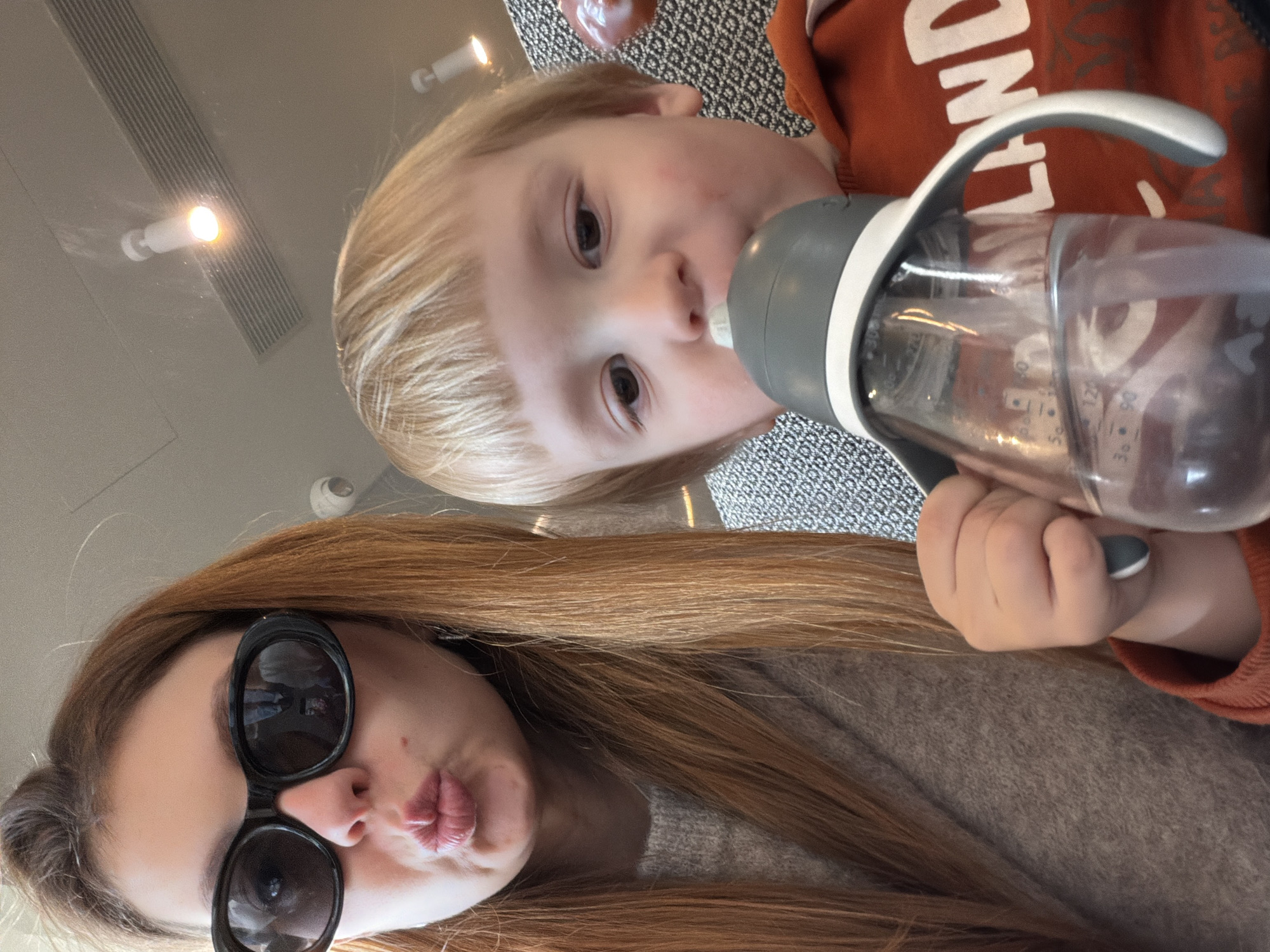 A selfie taken indoors features a young fair-skinned child drinking from a clear bottle with grey accents. The child has blonde hair and is wearing an orange top. In the foreground, a woman with long, brown hair and large sunglasses is pouting her lips towards the camera. Her shirt is a light brown color. The background includes a textured surface and a ceiling with visible light fixtures. The location appears to be an unspecified indoor setting in Unknown, Unknown.