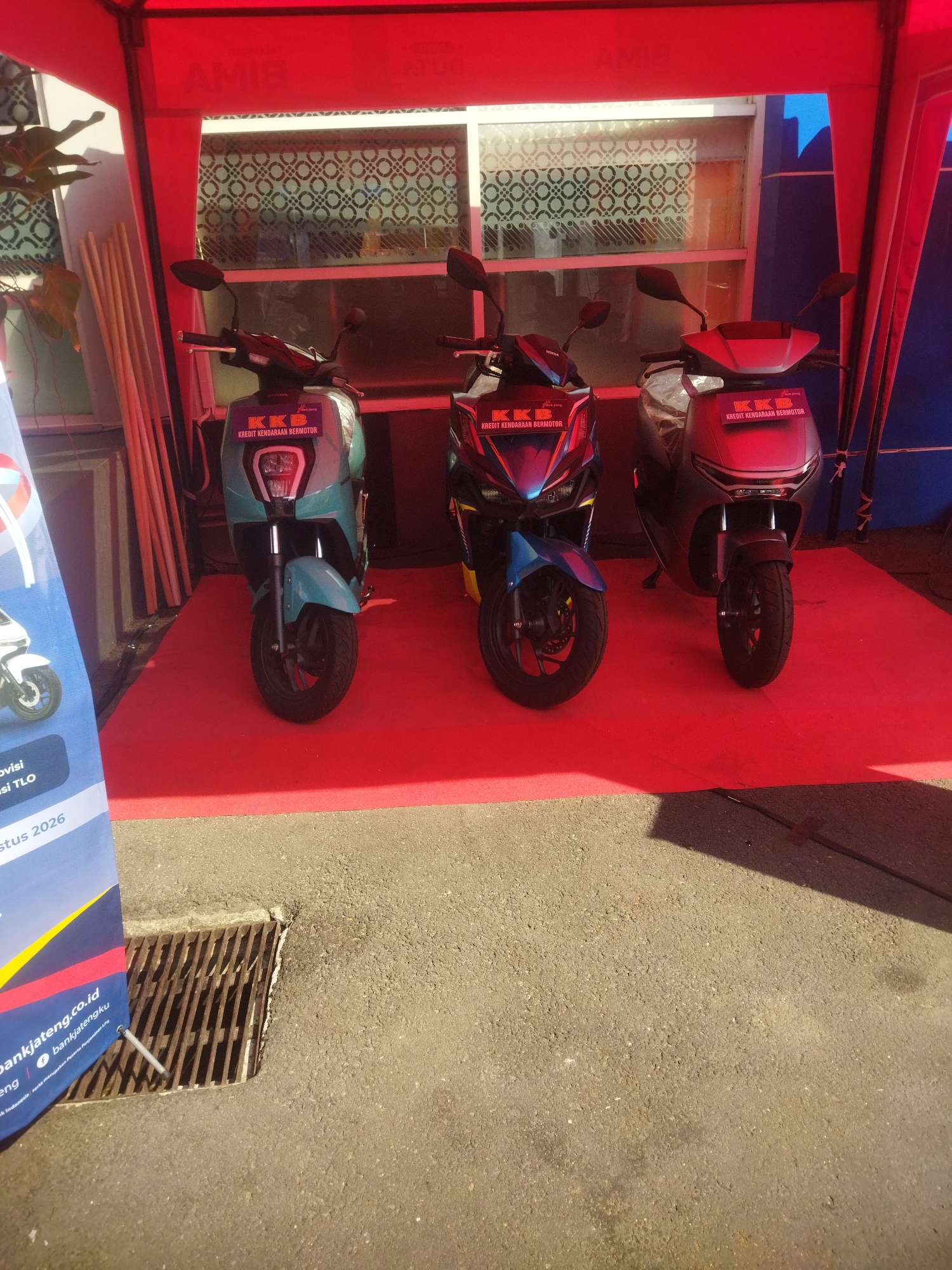 The image shows three electric scooters displayed under a red tent on a red carpet in what appears to be an outdoor setting. The scooters are lined up from left to right: a light teal scooter, a blue and black scooter, and a grey scooter. Each scooter has a "KKB KREDIT KENDARAAN BERMOTOR" sticker on its front. In the background, there is a window with patterned glass. To the left of the scooters, a blue banner with white text is partially visible, showing "bankjateng.co.id" and social media icons. The ground in front of the tent is asphalt with a metal grate. The scene suggests a promotional event or a dealership display for electric vehicles in Giripurwo, Indonesia.