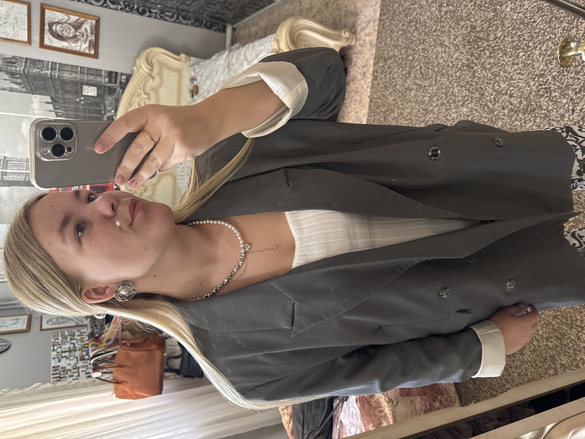 The image depicts a young woman taking a selfie in what appears to be a bedroom. She has blonde hair and is wearing a gray blazer over a white top, along with a pearl necklace and stud earrings. She is holding a smartphone in her right hand, with her fingers visible on the side of the phone. The background includes a bed with an ornate headboard, a mirror reflecting various objects including bags and clothes, and artwork on the wall. The location cannot be determined from the image.