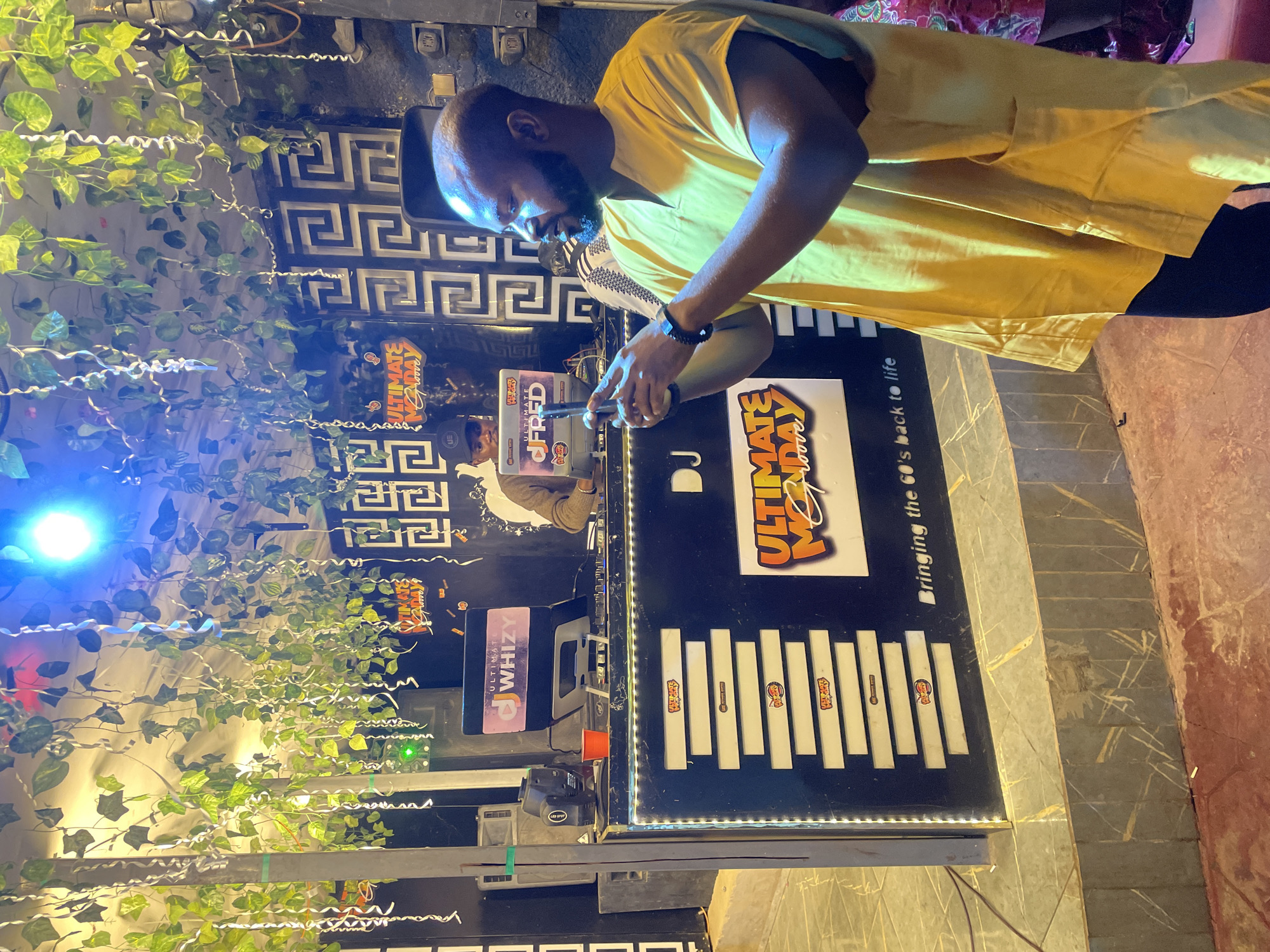 In Guzape, Nigeria, a man in a yellow shirt is holding a smartphone and appears to be recording a DJ setup. The DJ booth is decorated with lights and displays the name "ULTIMATE MONDAY GROOVE." There are plants and streamers hanging from the ceiling, suggesting a festive atmosphere. Two DJs are visible behind the booth, one working on the equipment and another looking at a laptop.