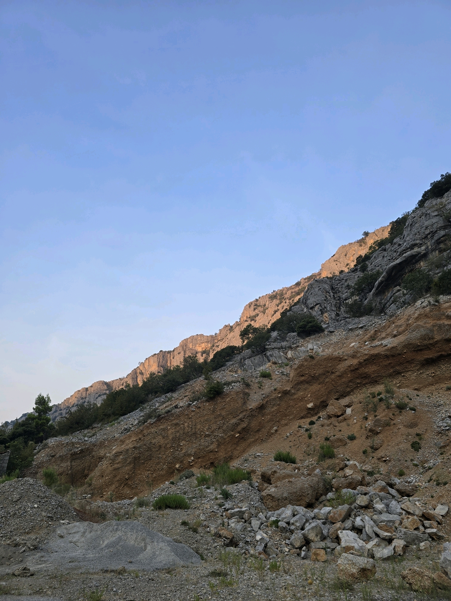 The image depicts a rugged, rocky mountainside under a clear, pale blue sky. The sun casts a warm, golden light on the upper slopes of the mountain, highlighting the textured rock faces and sparse vegetation. The lower part of the image shows a quarry or landslide area, with piles of gravel and large rocks scattered across the ground. A thin line of trees and bushes lines the base of the mountain, adding a touch of green to the otherwise earthy tones. The scene appears to be a natural landscape with evidence of human activity in the form of the quarry. Based on the query's context and the visual evidence, this scene is likely located in an area near Podgorica, Montenegro, possibly in a mountainous region or a designated quarry site within its vicinity, known for its limestone and rugged terrain.