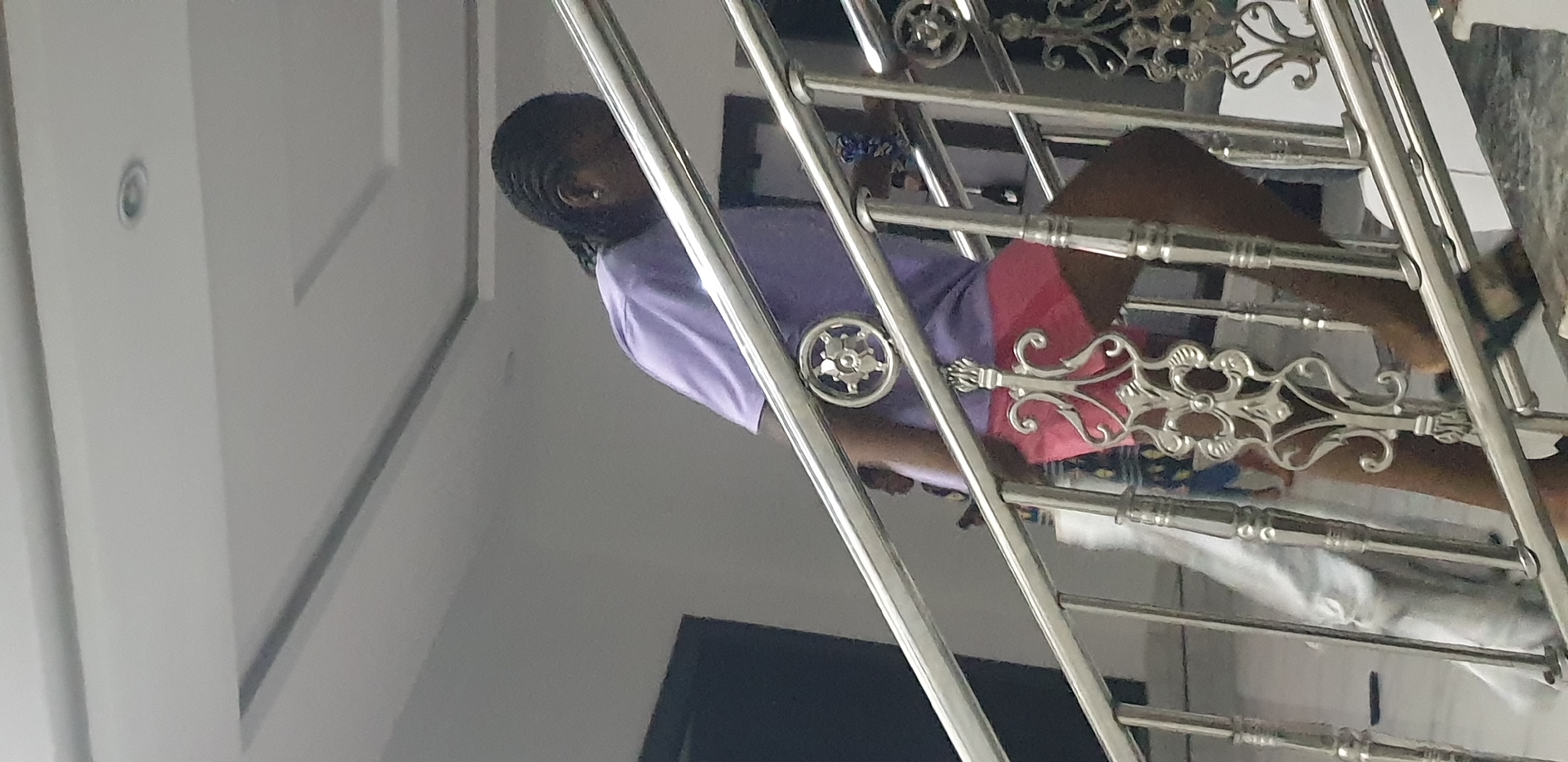 In Narayi, Nigeria, an interior scene depicts a person ascending a flight of stairs. The individual, wearing a light purple short-sleeved top and pink shorts, is positioned in the mid-ground, moving from left to right and upwards. Their dark hair is styled in braids, and a small, light-colored earring is visible in the left ear. Dark sandals or slippers are worn on the feet. A blue patterned item, possibly a scrunchie, is present on the right wrist. The person's head is slightly bowed, facing downwards.
The stairs are constructed with light-colored, possibly tiled or stone, treads and risers. A prominent silver-colored metallic railing with decorative scrollwork balusters and posts runs alongside the staircase. Circular floral motifs are present on some of the newel posts at the railing's top bar level. In the background, partially obscured by the railing, a second individual wearing patterned clothing is faintly visible at a lower level. The surrounding walls are painted in a light grey or white hue. The ceiling above is white with recessed lighting fixtures and decorative, multi-level false ceiling panels, some of which feature linear accents. Dark rectangular shapes, indicative of doorways or open thresholds, are visible in the background against the light walls.
