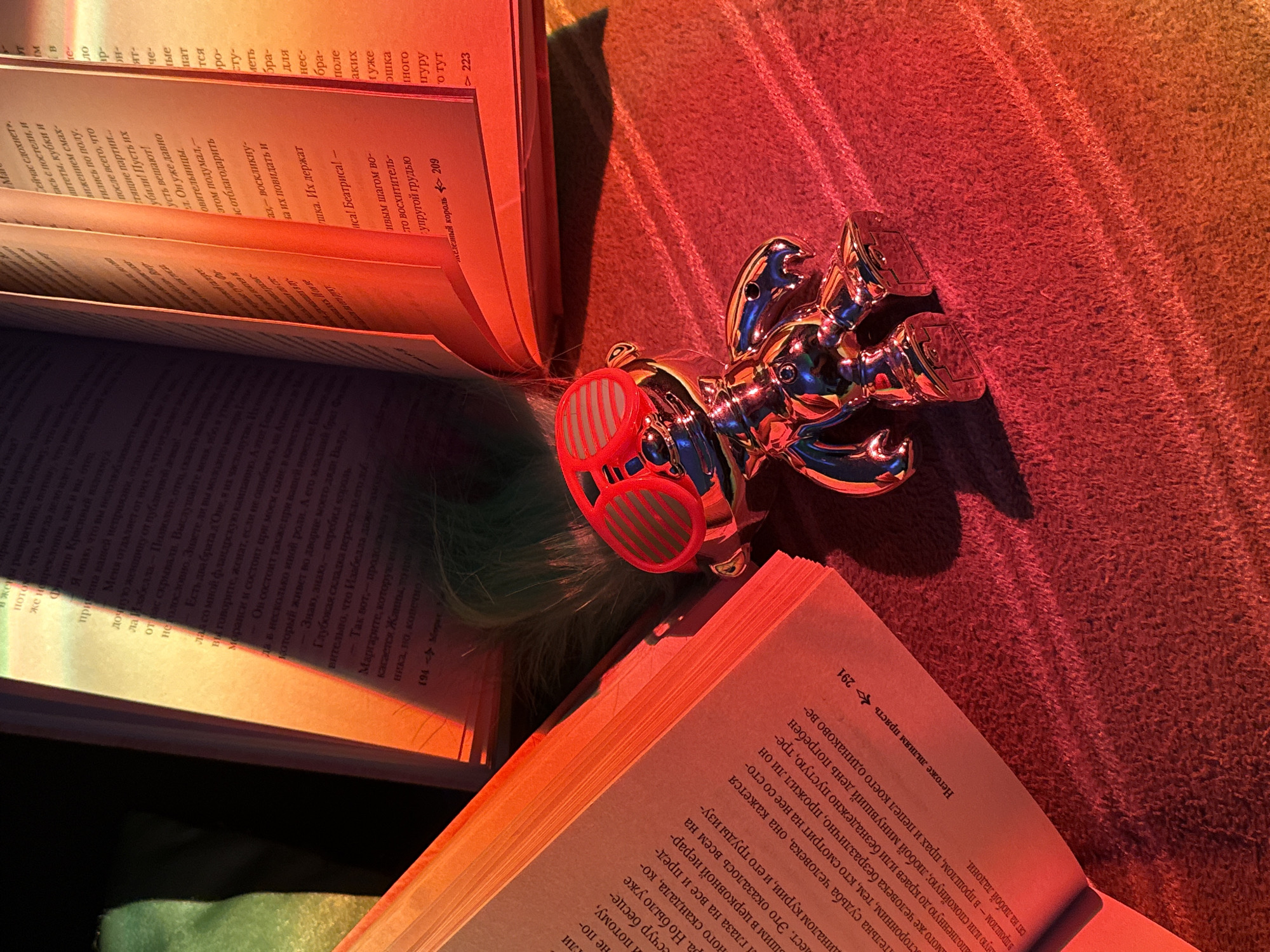 The image features an open book with Russian text, illuminated by a warm, orange light source. Another open book is stacked above it, and a chrome robot figurine with red, cartoonish glasses is resting on the pages. The background is a textured brown surface, possibly a blanket or rug. The lighting creates strong contrasts and highlights the objects, emphasizing the textures of the book pages and the metallic finish of the robot. The text in the books is not fully legible, but page numbers 291 and 209 are visible.