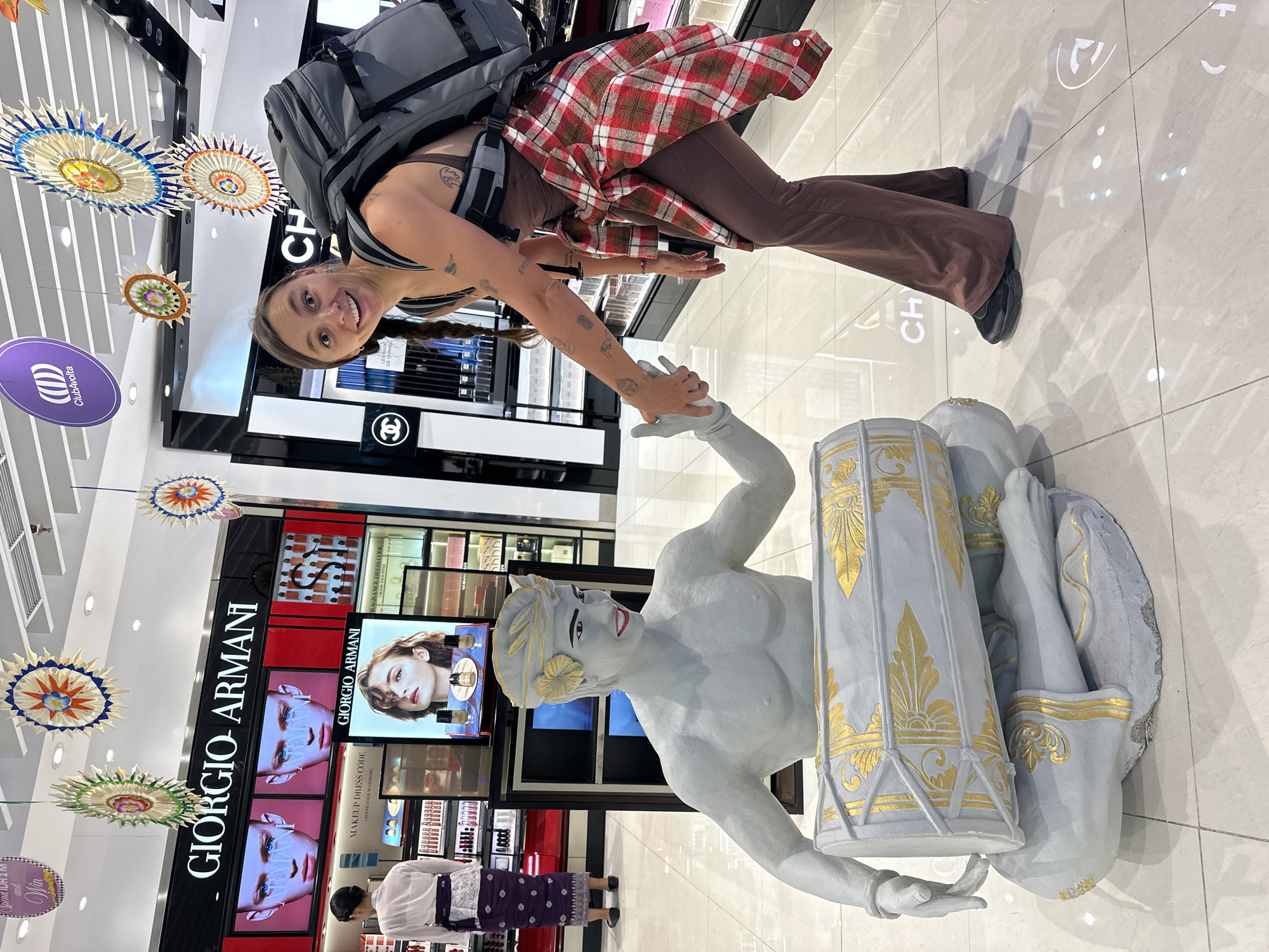 A brightly-lit retail space showcases Giorgio Armani and Chanel cosmetic and fragrance counters. Above, decorative paper sunbursts and the "ClubAvolta" logo hang from the ceiling. A woman with pigtails and a backpack stands holding the hand of a grey and gold statue of a deity. A sign is visible that says "Spend IDR 1 M and Win". Another person is browsing the Giorgio Armani display. The text "CH" is visible on the floor, as well as "EMFA". This scene is most likely taken in the city of Kuta, Indonesia.