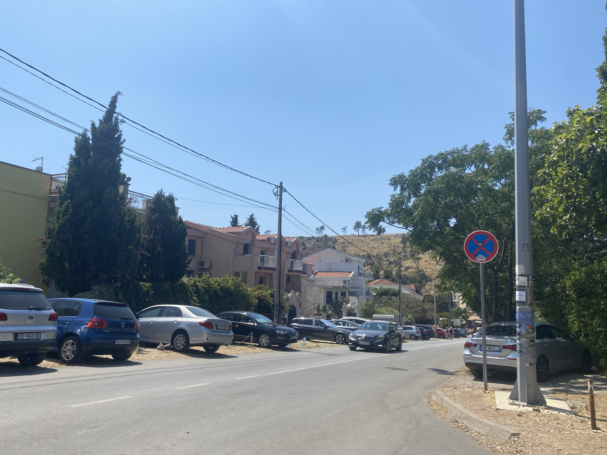 The image shows a street scene in Sutomore, Montenegro on a clear, sunny day. A row of parked cars lines the right side of the road. Among them is a silver sedan with license plate "MH512" visible. A tall, grey utility pole stands on the right side of the road, adorned with various stickers. A red and blue circular traffic sign prohibiting parking is affixed to the pole. Trees with dense green foliage are visible behind the parked cars and the utility pole. The street itself is paved, with a concrete curb and a gravelly shoulder. In the background, a hill with scattered trees and buildings can be seen. On the left side of the road, more parked cars are visible, including a silver Volkswagen, a blue Volkswagen Golf, and a silver sedan. A multi-story building with light-colored walls and balconies is situated behind these cars. The sky is a clear, bright blue.