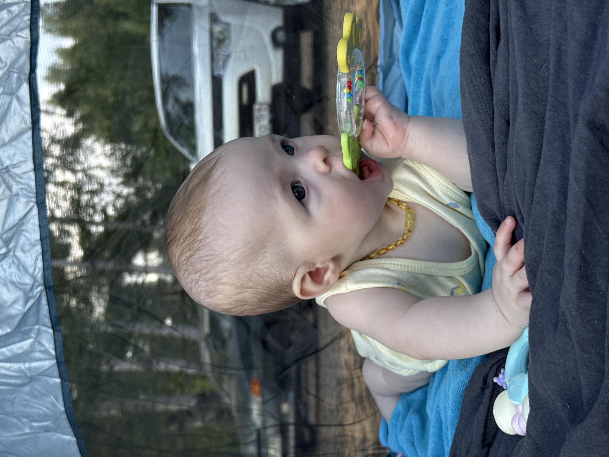 An infant is lying on their stomach, resting on a blue towel and a dark gray blanket. The infant is wearing a yellow top and an amber teething necklace. The infant is holding a green and clear rattle with colorful beads inside, with the rattle in its mouth. To the left of the infant, there are two other toys, one light purple and one light blue. In the background, two vehicles are visible behind a mesh screen, and beyond that, trees and foliage are apparent. The setting appears to be outdoors, possibly in or near Noviy, Russia, suggested by the presence of vehicles that are consistent with Russian license plates.