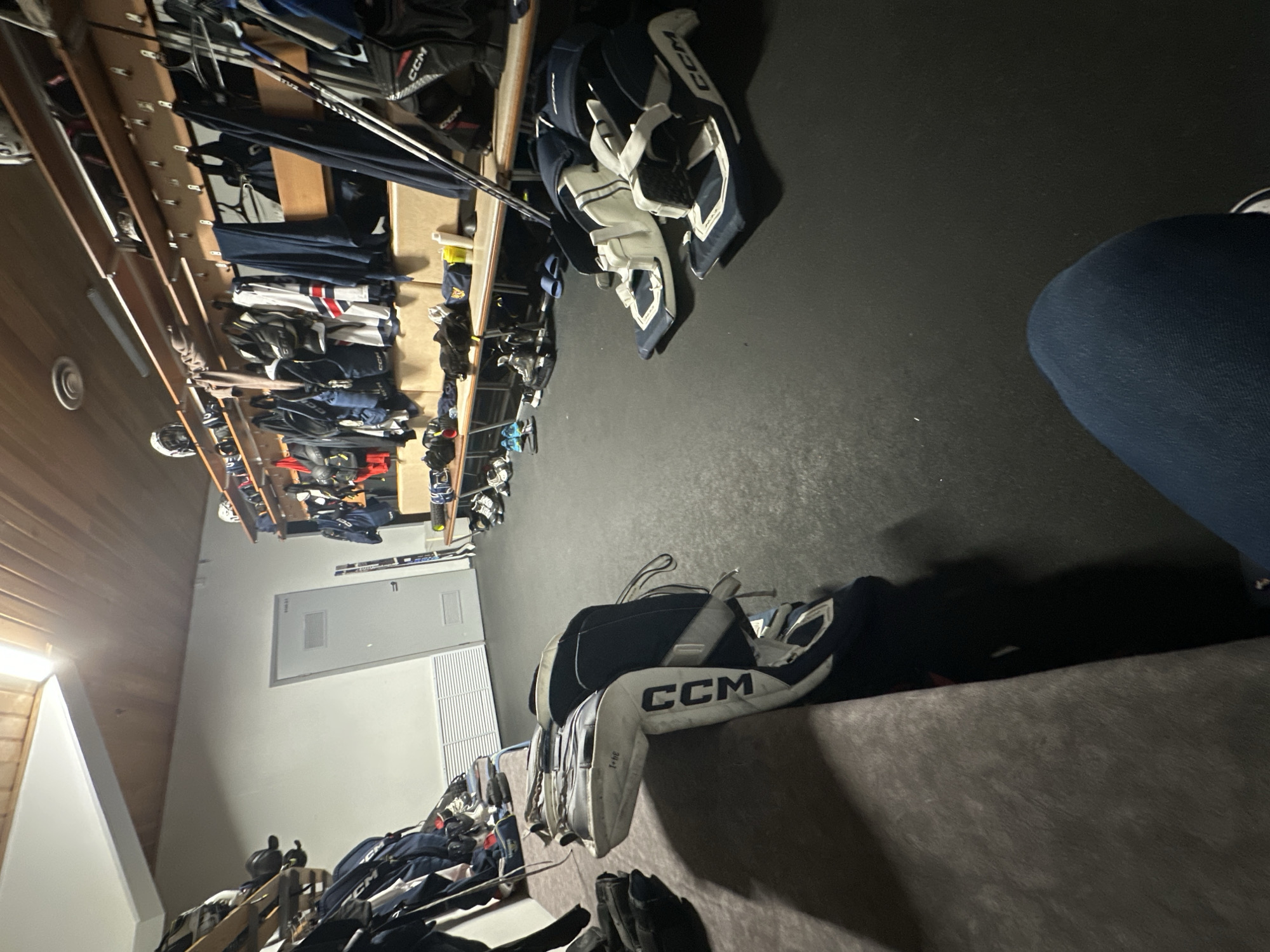 The image shows the interior of what appears to be a hockey dressing room in Unknown, Unknown. A long wooden rack system lines one wall, packed with hockey equipment including helmets, gloves, pads, and sticks. Hockey gear including a set of CCM goaltender pads are visible closer to the foreground. A gray door is partially visible along the wall. The floor is a dark matte color, possibly rubber or linoleum. Part of an individual wearing blue denim pants is present in the upper right of the image.
