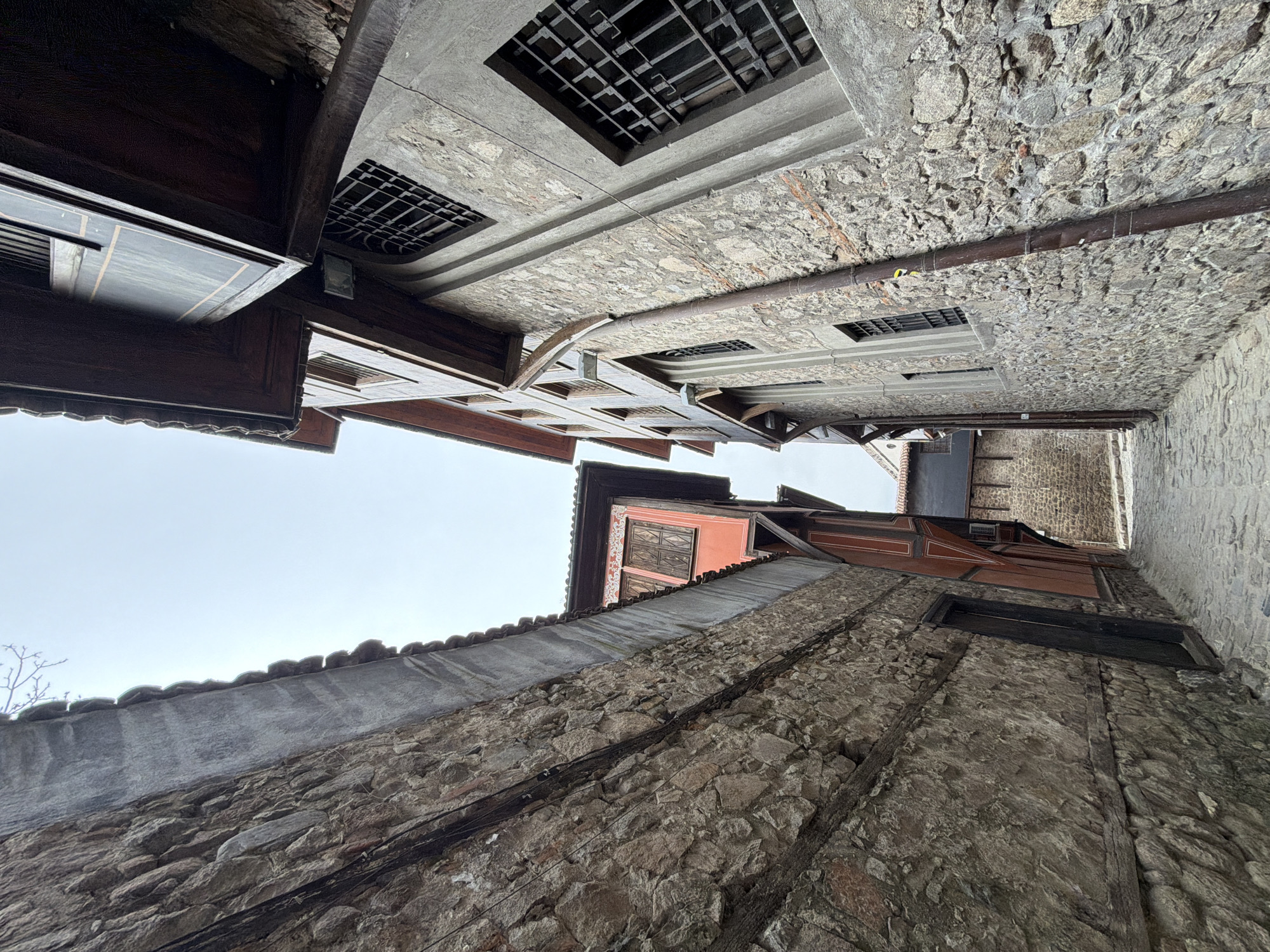 The image shows a narrow street in the Old Town of Plovdiv, Bulgaria, looking upwards at the buildings lining the street. The buildings are made of stone and wood, with some having visible windows and tiled roofs. The sky is overcast. A metal pipe runs along the wall. The architectural style is characteristic of the historic area, with a mix of stone facades and wooden support beams.