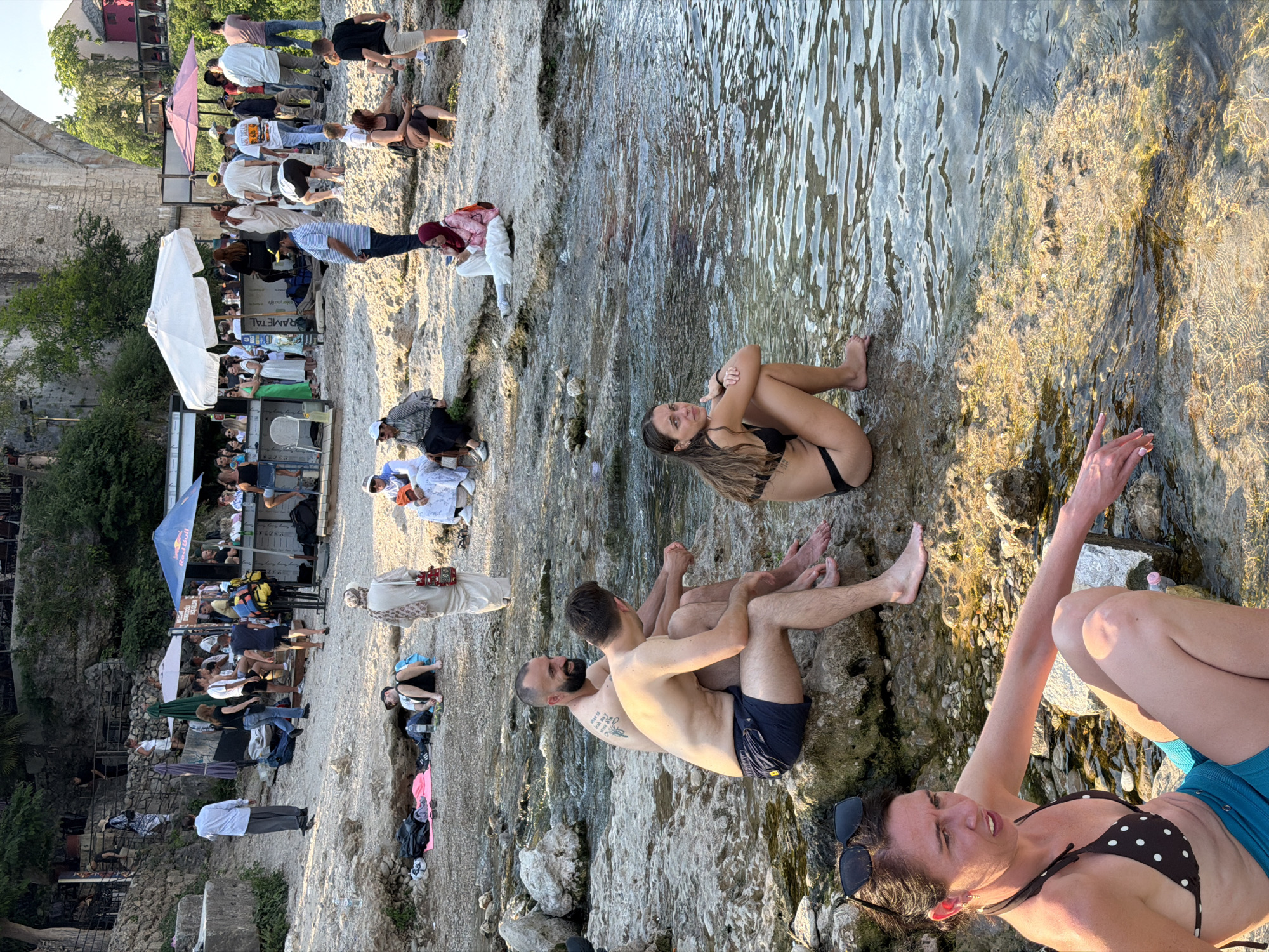 The image captures a lively scene along the banks of the Neretva River in Mostar, Bosnia and Herzegovina, likely near the iconic Stari Most (Old Bridge). In the foreground, several individuals are enjoying the water's edge. A woman in a polka dot bikini top and turquoise shorts sits on rocks, her hand extended towards the water. To her left, a man with a beard and tattoos, and another man sit together on the rocks. Further back, a woman in a black bikini is also seated on the rocks, looking towards the river.
The middle ground of the image reveals a bustling area with people gathered around cafes and stalls set up on the rocky embankment. Some are seated, others are standing, and there's a general atmosphere of social activity. In the background, the imposing structure of the Stari Most bridge is visible, arching majestically over the river. Lush green vegetation and the natural rocky landscape contribute to the scenic beauty of the location. The overall impression is one of a popular tourist spot, with people relaxing and enjoying the river and the historic surroundings.