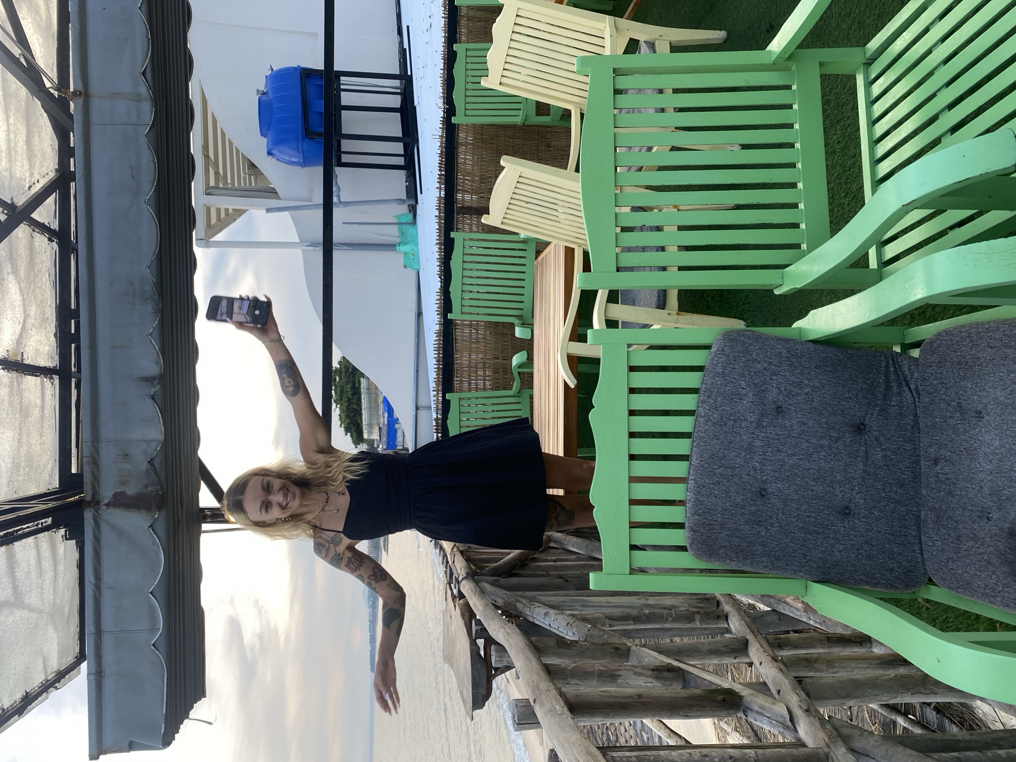 A young adult Caucasian female is standing on a wooden balcony overlooking a beach and the ocean. She is wearing a black dress and has tattoos on her arms. She is holding a smartphone in her right hand, angled towards herself, suggesting she is taking a selfie or recording a video. Her left arm is extended out to her side, and she is smiling at the camera. In the foreground, there are several green wooden chairs with gray cushions, and a wooden table with wooden chairs. The balcony is constructed of rough-hewn wooden planks and railings. The background shows the ocean, a beach with some debris, and a white building with a blue water tank on its roof. The sky is overcast with some clouds. The location is identified as Phu Thuy Ward, Vietnam.