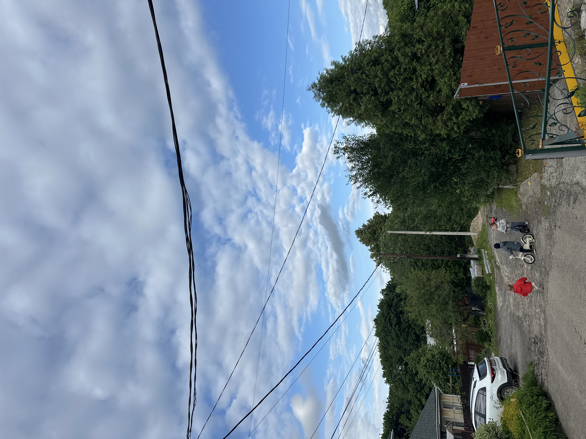 The image shows a view looking down a residential street in Nizhny Novgorod, Russia. The sky is blue with scattered white clouds, and power lines stretch across the frame. On the right side of the image, there is a house with a dark roof and light siding. Further down the street, there is a white car parked. In the center of the image, two children are riding bicycles on the road. A third child is walking toward them wearing a red jacket. The road appears to be unpaved or gravel. On the right side of the road, there is a house and some trees. A fenced area can also be seen to the right.