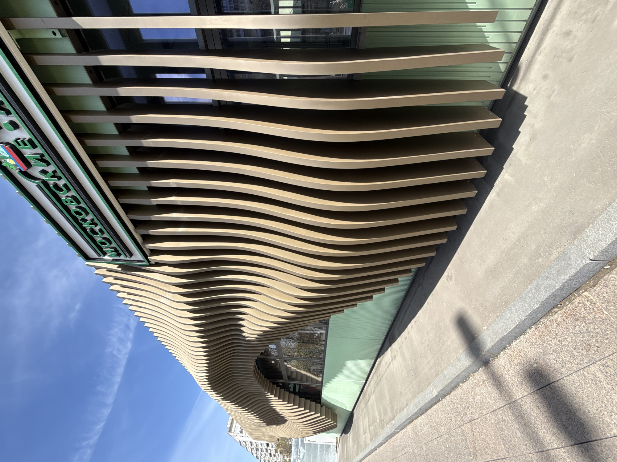 The image shows the facade of the "Cross Time" cafe located in Moscow, Russia. The facade is composed of horizontally-aligned, undulating wooden panels. The lower portion of the building features windows. A sign with green lettering and trim is visible on the left side of the structure. The sky is blue with a few light clouds. A sidewalk and pavement are visible in the foreground, with a faint shadow of a person. A tall, white apartment building can be seen in the background.
