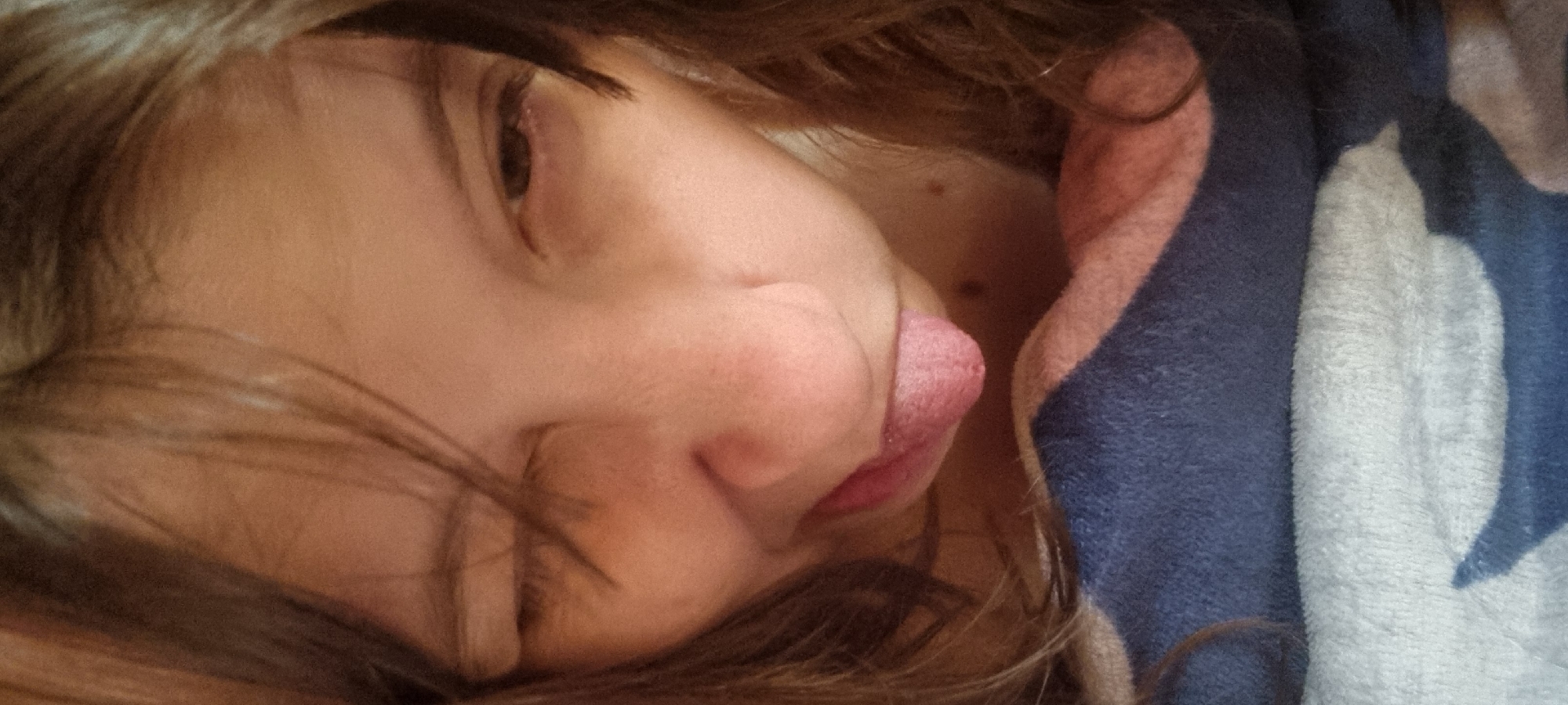 A close-up, horizontally-oriented photograph shows a child lying down. The child has brown hair, closed eyes, and a fair complexion. Their tongue is sticking out slightly. The child appears to be resting against a soft, patterned blanket, which has shades of blue, white, and pink. The exact location within Dunnae-myeon, South Korea, is indiscernible from the visual data.