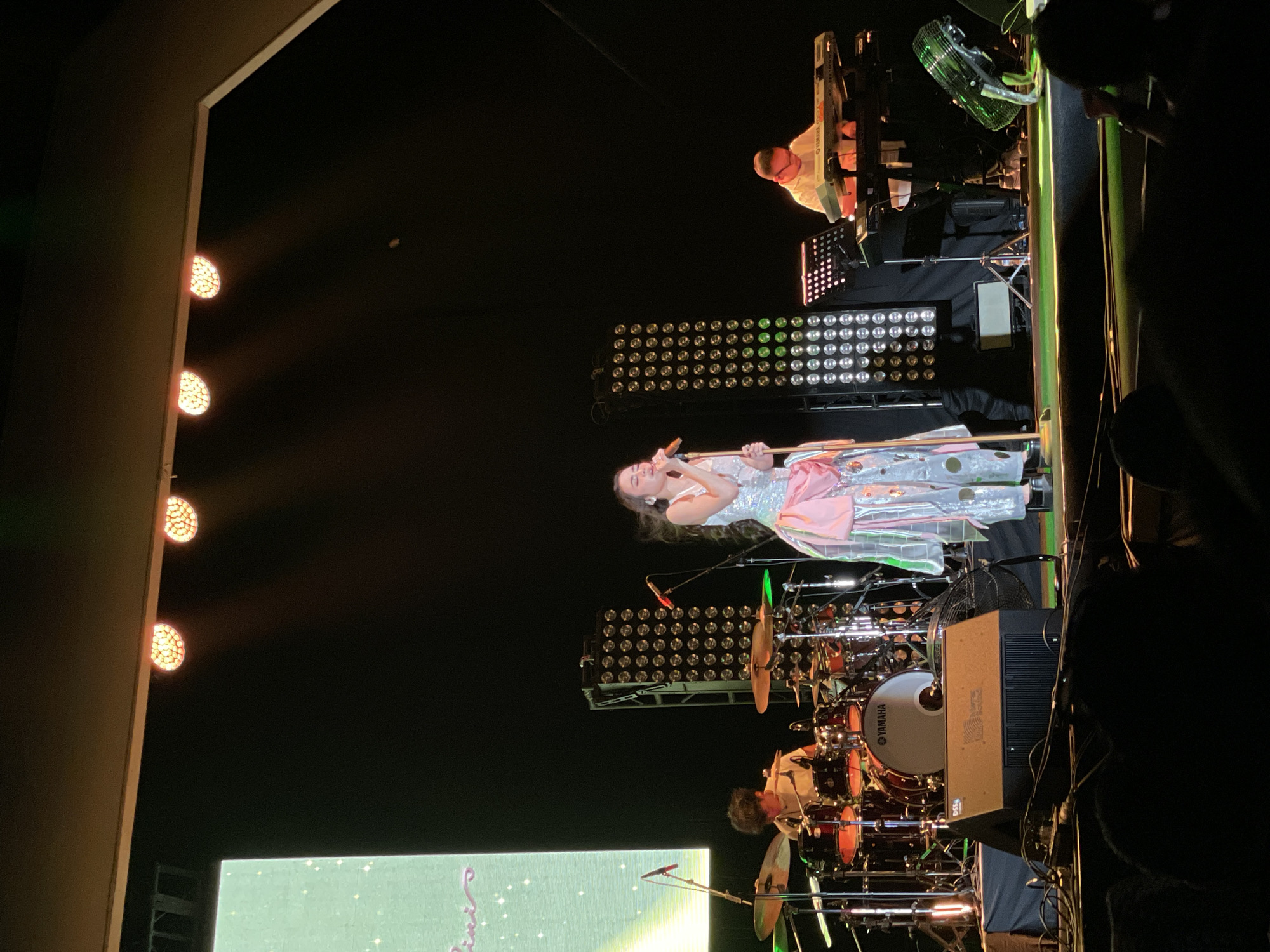 The image depicts a musical performance on stage, presumably at a concert venue, though the specific location in Salembaran, Indonesia is not discernible from the image. A woman dressed in a sparkly, light-colored dress with a pink sash is singing into a microphone. She is positioned center stage. Behind her are numerous spotlights and a drum set with a drummer visible. Above and behind the singer, a keyboard player is visible, playing on a Yamaha keyboard. To the far right edge of the photo, there appears to be a cooling fan. The left edge of the image shows a building or stage structure with orange lights. A large screen is visible in the left foreground, projecting a green-toned image. The foreground shows indistinct silhouettes of an audience.
