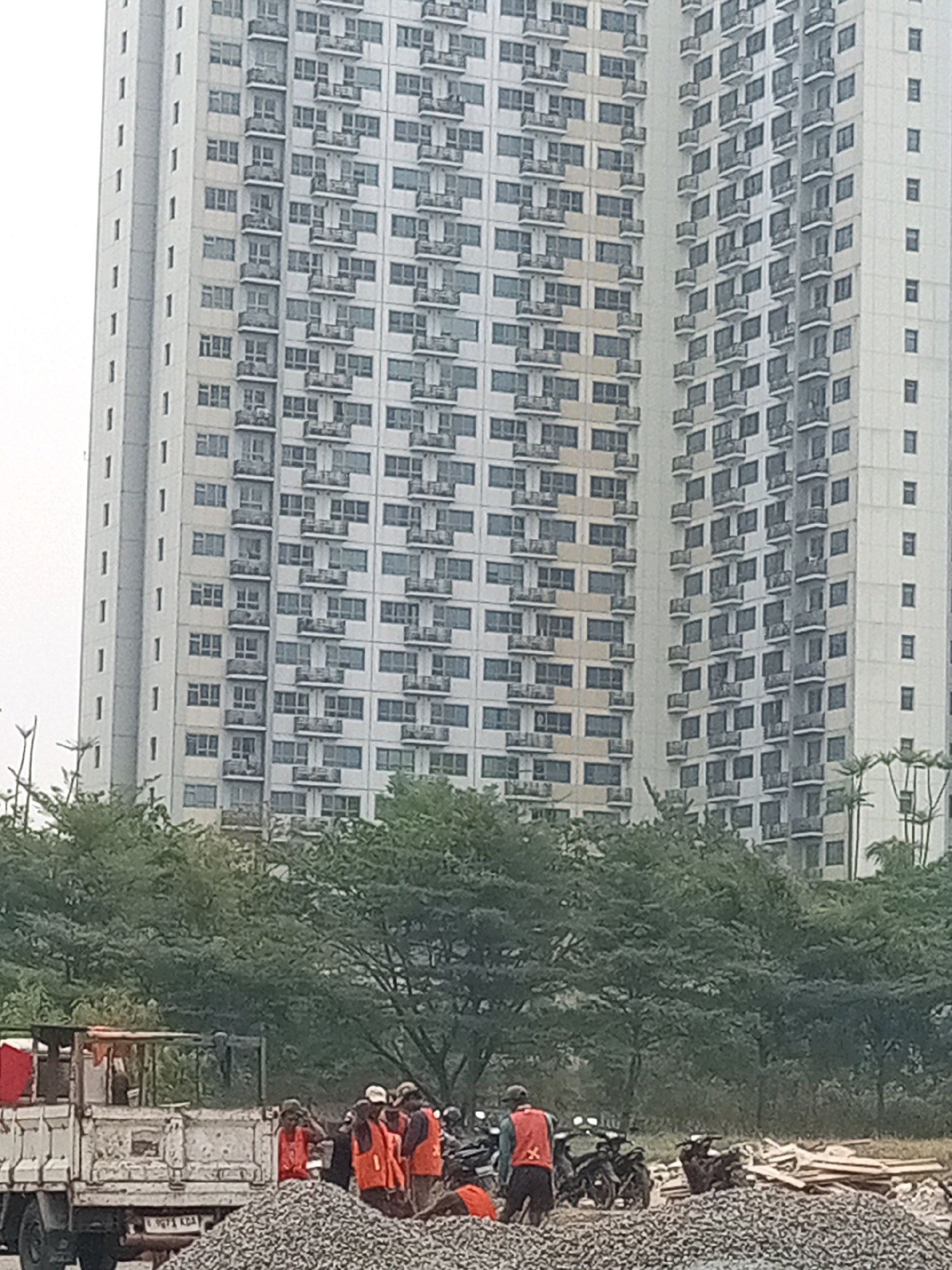 The image depicts a construction site in Bekasi, Indonesia, with two tall, multi-story apartment buildings in the background. In the foreground, a white flatbed truck is parked next to a large pile of gravel. Several construction workers, dressed in orange vests and hard hats, are present at the site. Some appear to be engaged in work, while others are standing around the truck and the gravel pile. Motorcycles are also visible near the workers, suggesting a form of transportation for them. The environment appears to be an urban setting, with dense greenery surrounding the construction area, indicating it might be located in a developing part of the city. The sky is overcast, suggesting it might be a cloudy day.