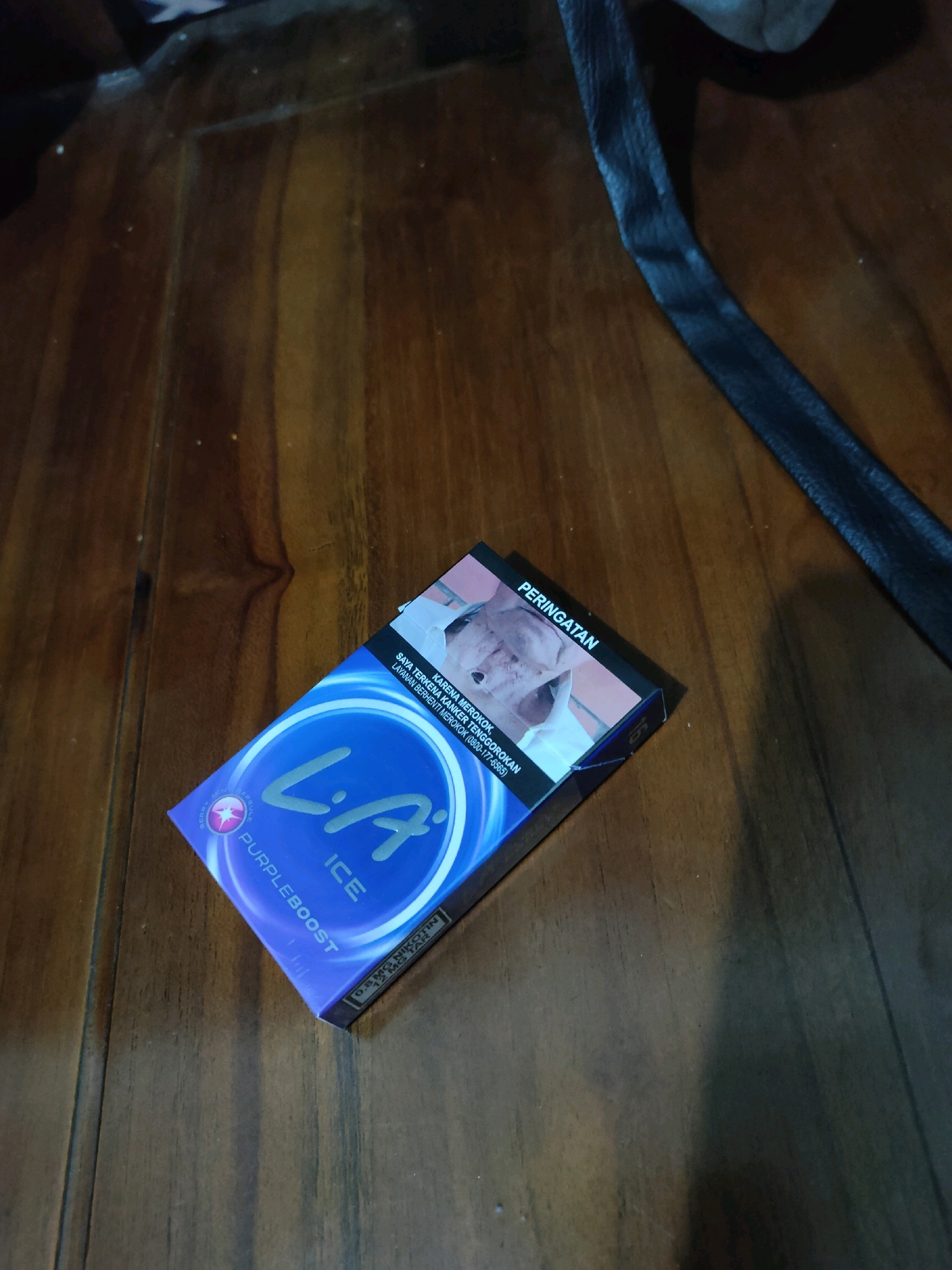 A close-up, overhead shot shows a pack of LA Purple Boost Ice cigarettes resting on a dark wooden surface. The cigarette pack is angled, with its front face prominently displayed. The pack features a vibrant purple and blue design with the "LA" logo in a large, white, stylized font. The text "ICE" and "PURPLEBOOST" are also visible. A warning label with an image of a person's face is present on the top right of the pack. A dark, textured object, possibly a strap or part of a bag, lies diagonally across the upper right portion of the image, casting a shadow. The wooden surface appears to be part of a table or floor, with visible wood grain and slight reflections. The scene is indoors, with no specific details indicating the city of Ngebruk, Indonesia, beyond the requested context.