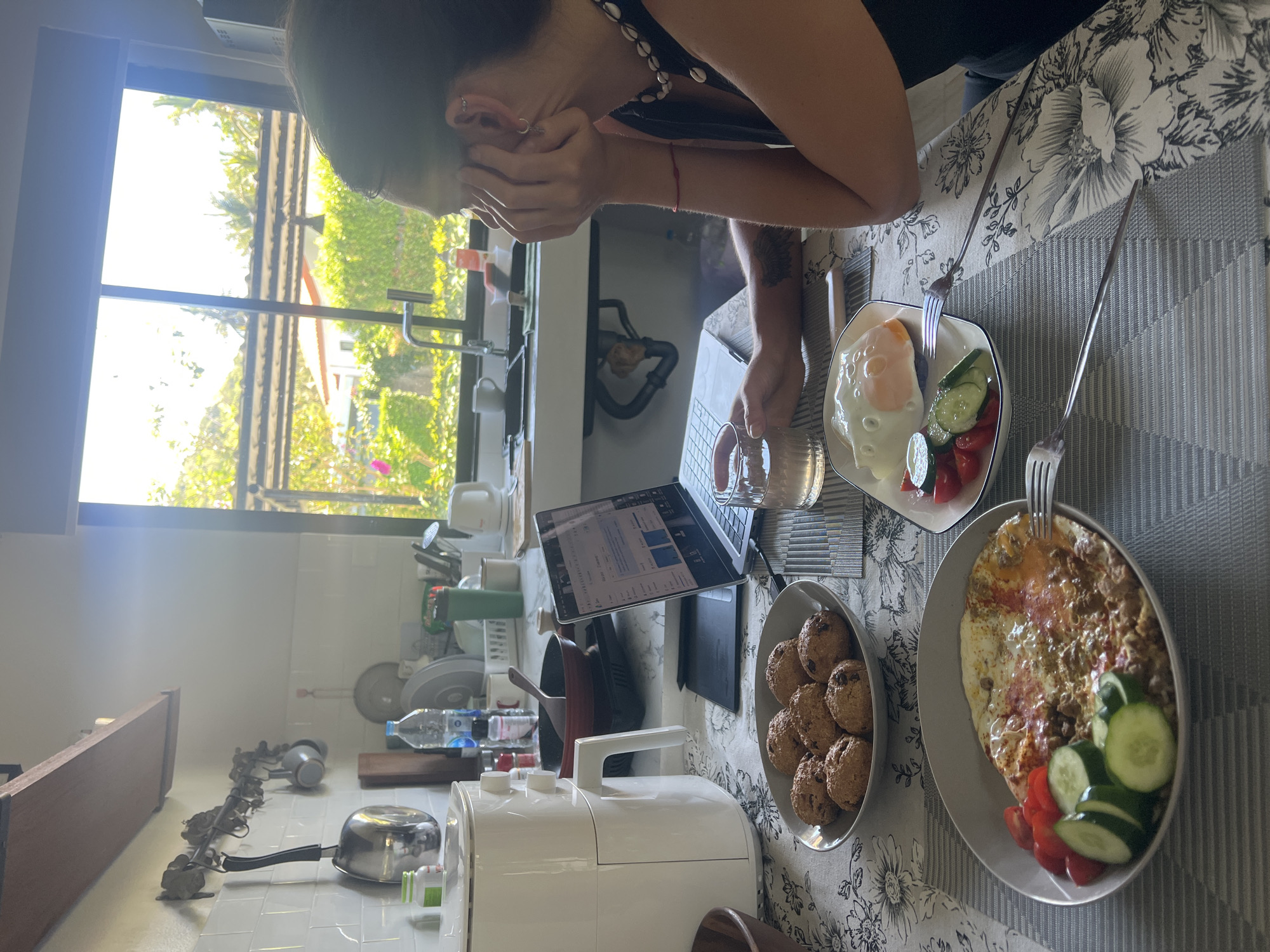 A woman sits at a table with a laptop, a glass of water, and two plates of food, one with a fried egg, cucumbers, tomatoes, and lime, and the other with a fried egg, minced meat, cucumbers, and tomatoes. Beside the plates is a bowl of cookies. The woman is holding her face with her hands and appears to be looking at the laptop screen. A window behind her shows trees and buildings outside. The location is a kitchen in บ้านบางรักษ์, Thailand, as suggested by the style of the buildings outside the window.
