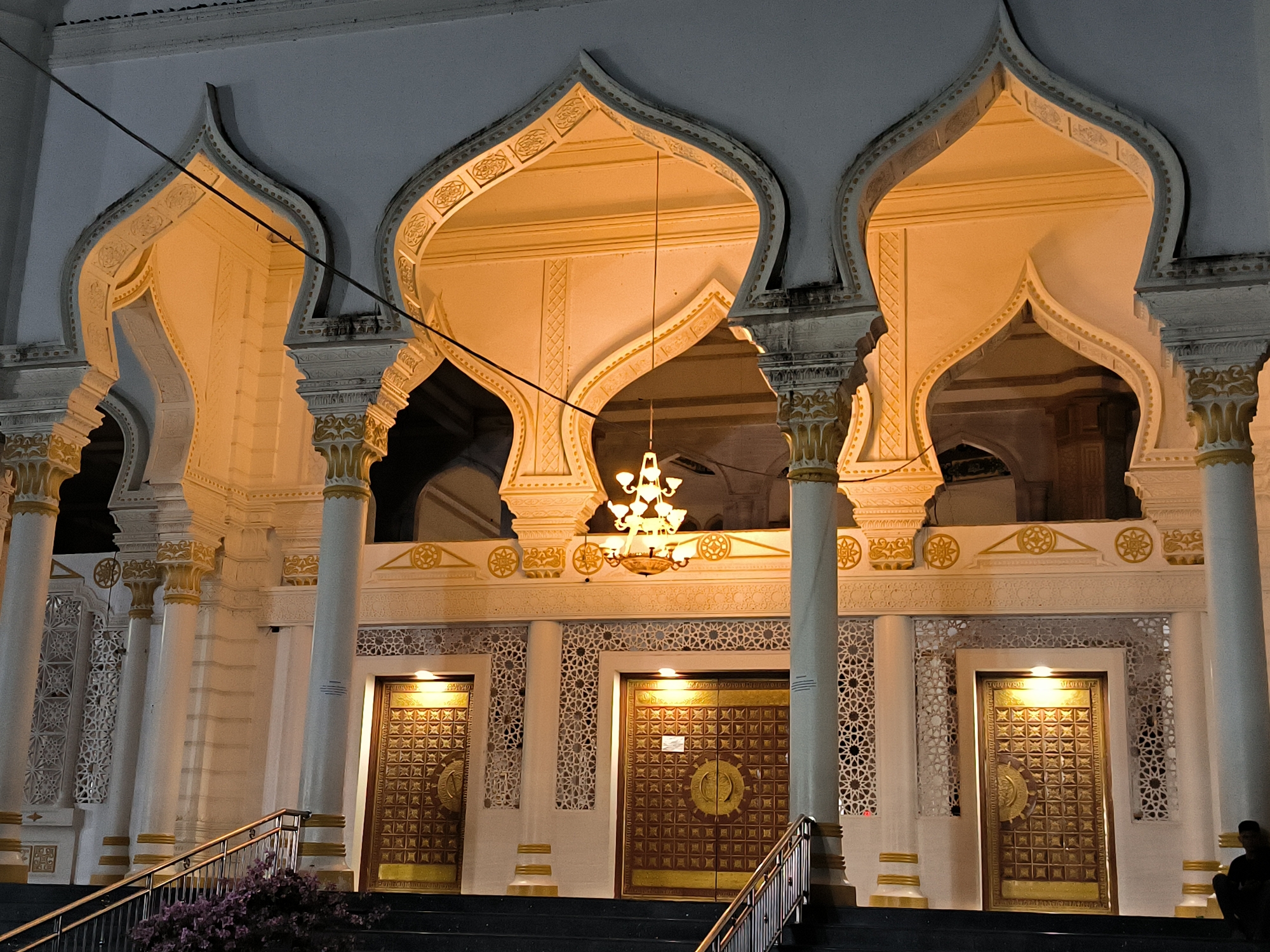 A low-angle, nighttime shot depicts the main entrance of a building featuring Islamic architectural elements. The architecture is painted primarily in white with gold ornamentation. Three large arched openings are visible above three heavy, dark-wood doors, each adorned with metallic fittings. A chandelier hangs centrally in the middle arched opening. Columns flank each doorway. There are stairs leading up to the doors with metal railings. A small bush with purple flowers is at the bottom of the stairs on the left. Wires run across the top of the image. A figure is sitting on the steps on the right. The city is Kotapantonlabu, Indonesia.