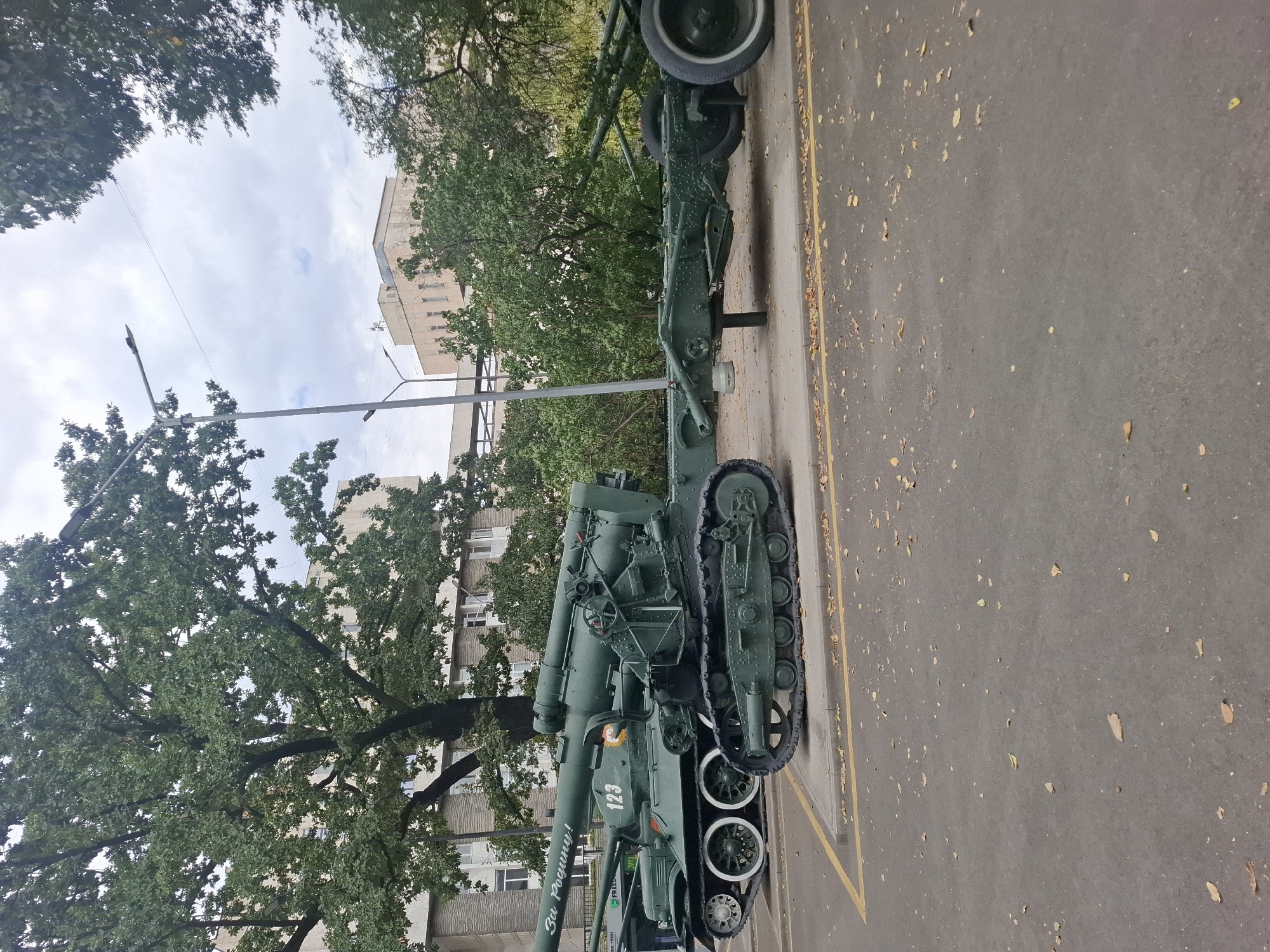 The image depicts two military vehicles, a tank and a towed artillery piece, parked on an asphalt surface with yellow and white lines marking a border. The tank, painted olive green, has the number "123" on its side and an inscription that appears to read "За Родину!" (For the Motherland!). It is positioned to the left of the artillery piece. The artillery piece is also olive green and features tracks and wheels. Both vehicles are stationary, and no people are visible. The environment includes a large tree with lush green foliage, several buildings in the background, streetlights, and scattered fallen leaves on the ground, suggesting it may be autumn. The overall scene appears to be an outdoor exhibit or memorial, potentially in a public space within Almaty, Kazakhstan, given the context.