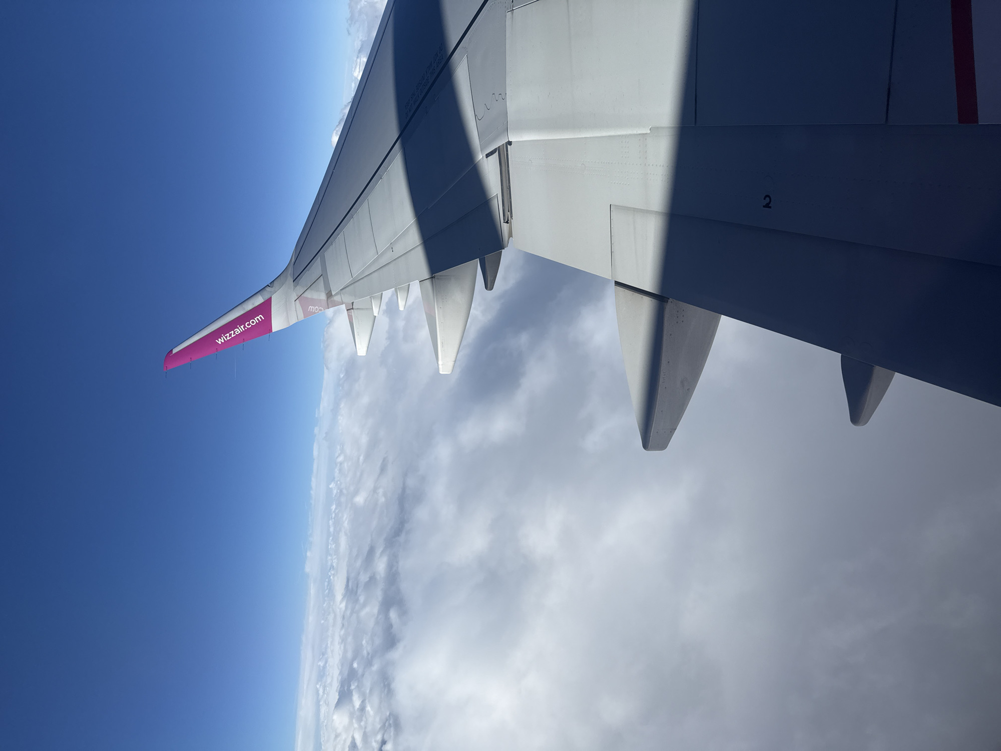 This media file captures an aerial view from an airplane window, dominated by a large portion of the aircraft's wing against a backdrop of sky and clouds. No people are visible in the image.
The main subject is the gray-white wing of a passenger plane, positioned on the right and extending towards the center of the frame. The wing displays various aerodynamic features, including control surfaces, panels, and rivets. A distinctive bright magenta section on the wingtip features the white text "wizzair.com," identifying the airline as Wizz Air.
Above and to the left of the wing, a wide expanse of clear, deep blue sky is visible, suggesting a bright, sunny daytime environment at high altitude. Below the wing and across the right side of the frame, a textured layer of white and gray clouds stretches out, indicative of fair weather conditions. Shadows from the wing are cast onto the cloud layer and parts of the wing itself, emphasizing the strong sunlight.
Visible text includes "wizzair.com" prominently on the wingtip. Other texts partially discernible on the wing surface are "DO NOT WALK OUTSIDE THIS AREA" and the number "2". A reversed "COW" is also faintly visible, possibly a reflection or text from the underside.
The image is rotated 90 degrees counter-clockwise, giving the impression of a vertical horizon, with the blue sky on the left and clouds on the right. The airplane is clearly in flight during the day. While the general location context is Künersberg, Germany, no specific ground features are identifiable from this high-altitude view above the clouds.