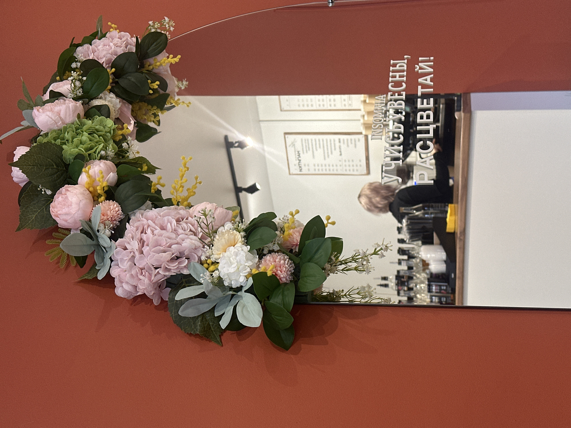 A close-up shot shows a mirror, partially adorned with a floral arrangement, against a solid terracotta background. The floral arrangement, consisting of pink hydrangeas, roses, and greenery, is affixed to the mirror's edge. Reflected in the mirror is the interior of a café or restaurant. A barista is seen behind the counter, surrounded by bottles and equipment. Menu boards can be seen on the wall behind the counter. The text "INSOMNIA УЧИСЬ УВЕСНЫ, РАСЦВЕТАЙ!" is written on the mirror. Based on the text and the reflection, the location is Chaikovsky, Russia.