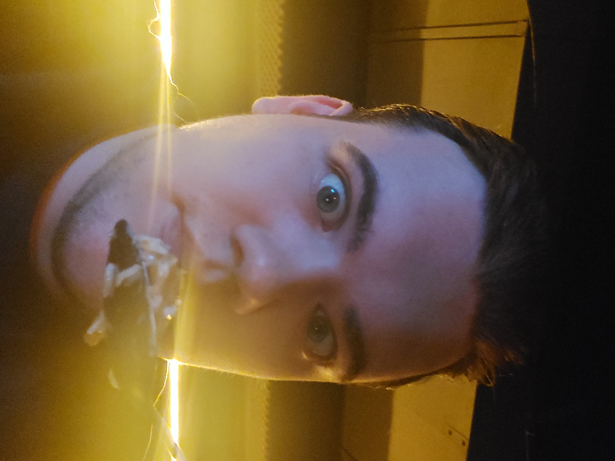 A close-up, rotated view depicts a person's face with light-colored skin, dark hair, and noticeable eyebrows. The eyes are wide open, looking up and to the right. The person's mouth is open and something is in it, obscured by light and blur. Part of the person's ear is visible at the top of the frame.
The background consists of a blurred mix of yellow and dark tones. Some linear light streaks are visible, suggesting a light source or reflection. The exact location cannot be determined without additional context or information. The image provides no indication of Tashkent, Uzbekistan.