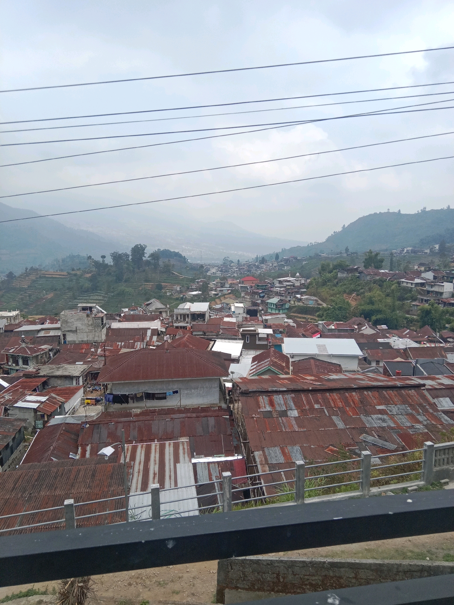 The image depicts a dense residential area in Surengede, Indonesia, characterized by numerous houses with predominantly corrugated tin roofs, many of which show signs of rust and weathering. The houses are clustered closely together, forming a hillside settlement with varying roof heights and materials. The architecture appears to be simple, with many structures made of concrete and brick.
In the foreground, a dark railing runs horizontally across the bottom of the frame, suggesting the vantage point is from an elevated structure or a balcony. Beyond the railing, a patch of dry, dusty ground is visible.
The middle ground is dominated by the rooftops of the houses. Some roofs are a uniform reddish-brown, while others are a patchwork of different colored and weathered tin sheets, indicating repairs or replacements over time. Clotheslines with laundry are visible hanging between some houses, a common sight in residential areas.
In the background, the settlement extends up a verdant hillside. Terraced fields are visible on the slopes, indicating agricultural activity in the area. Further in the distance, a hazy mountain range forms the backdrop to the scene, with a light-colored sky suggesting overcast conditions or early morning/late afternoon light. Power lines are strung across the upper part of the image, adding to the sense of a lived-in, functional urban or semi-urban environment. There are no clearly discernible people or specific actions captured in the frame, contributing to a general scene of everyday life in a hillside town.