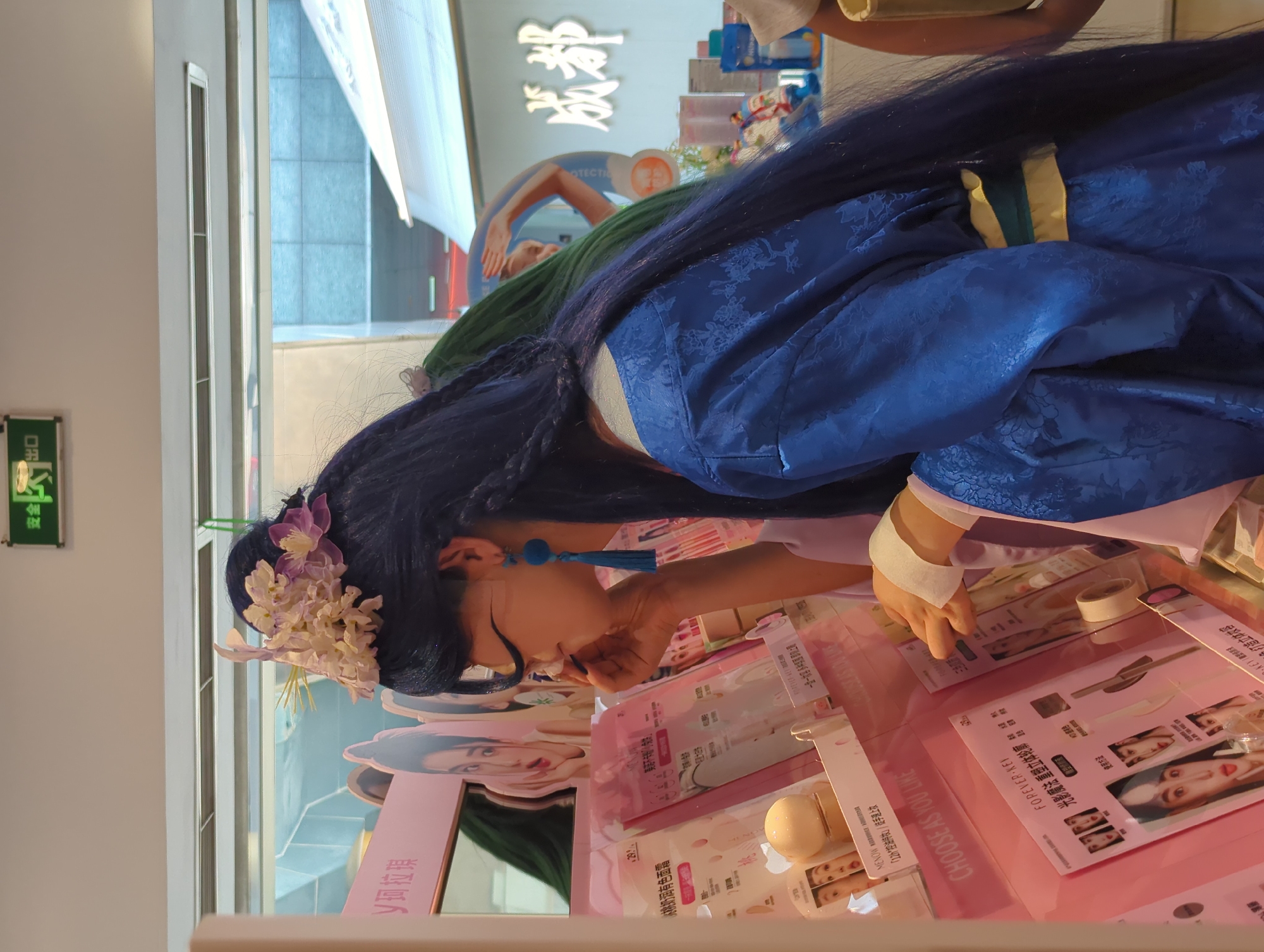 In a retail store in Qingyang District, China, a person dressed in a blue robe with a floral pattern and a long blue wig adorned with white and purple flowers is examining makeup products. The person is wearing large blue tassel earrings. They are leaning towards a display rack filled with pink packaging of various cosmetic items, including what appears to be foundation or cushion makeup. A hand with a white bandage on the wrist is extended towards the products. Behind the person, a sign with Chinese characters is visible. Further in the background, another person with green hair can be seen, and a large sign with "成都" (Chengdu) is illuminated. The overall environment suggests a beauty or cosmetics retail setting.