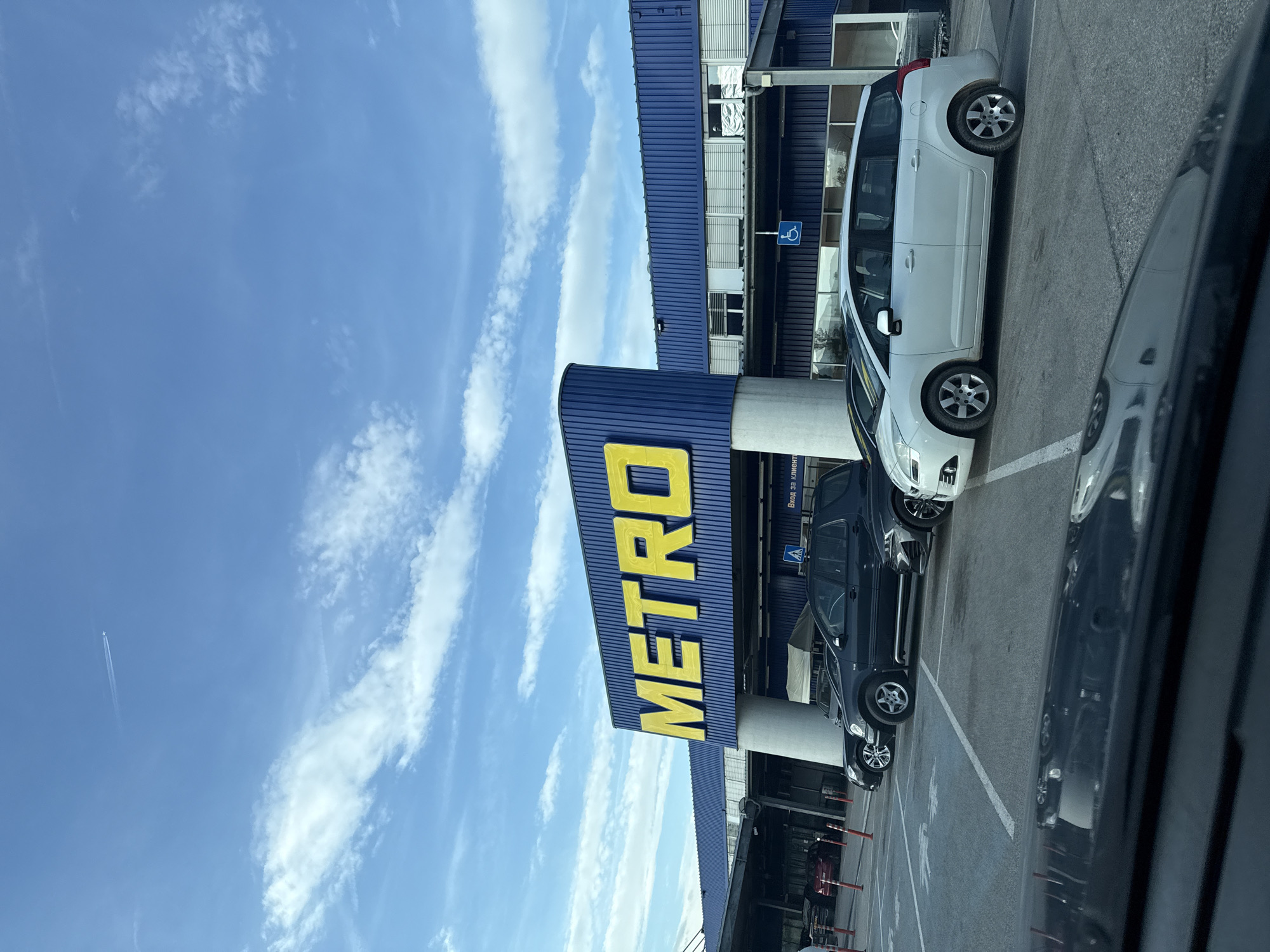 The image shows the exterior of a METRO Cash & Carry store, likely in Stara Zagora, Bulgaria (as the sign says "Вход За клиенти"), as the store's signage is prominently visible. The building has a dark blue facade with a large yellow "METRO" sign. The sky is blue with scattered white clouds. Several cars are parked in a parking lot in front of the building. A white car and a dark car are clearly visible. A sign with a wheelchair symbol is present near the entrance.