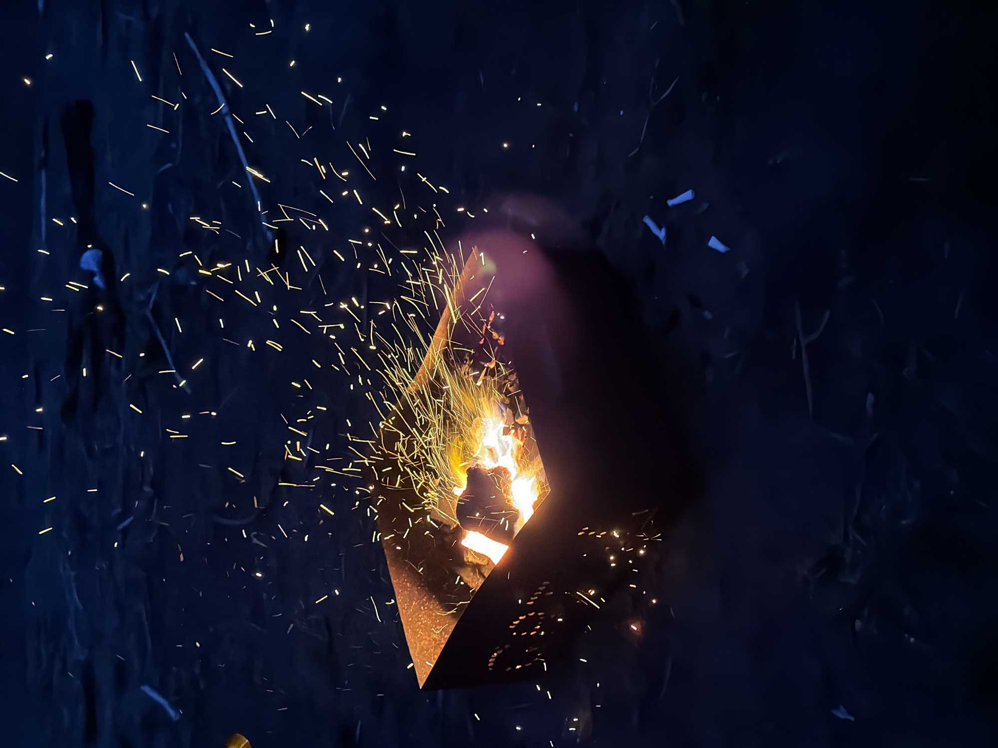 A fire is burning in a metal fire pit, with sparks flying. The surrounding environment is dark. No people are visible. The location is given as Nikolo-Beryozovka, Russia.
