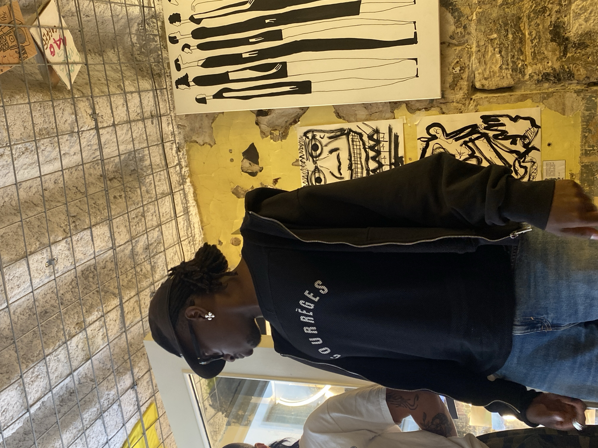 The image depicts a man wearing a black "Courrèges" jacket, a cap, and glasses. He's indoors, likely in a gallery or art space, as suggested by the artwork on the weathered yellow wall behind him. The artwork includes a black and white linear drawing of figures and some abstract sketches. A metal grid is visible on the left side. Another person is partially visible in the bottom part of the image. The scene is possibly set in the city of Paris, France, based on the jacket branding and the urban aesthetic, although there is no definitive location marker within the frame.