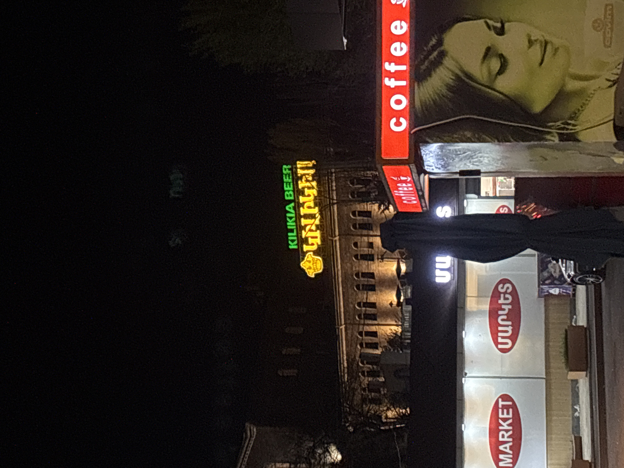 The image shows a night scene in Yerevan, Armenia, with storefronts visible. "KILIKIA BEER" is illuminated in green neon. Next to it is a "COFFEE" sign in red neon. Adjacent is a sign with a person's face. A market sign is visible that reads "MARKET" and an Armenian word.