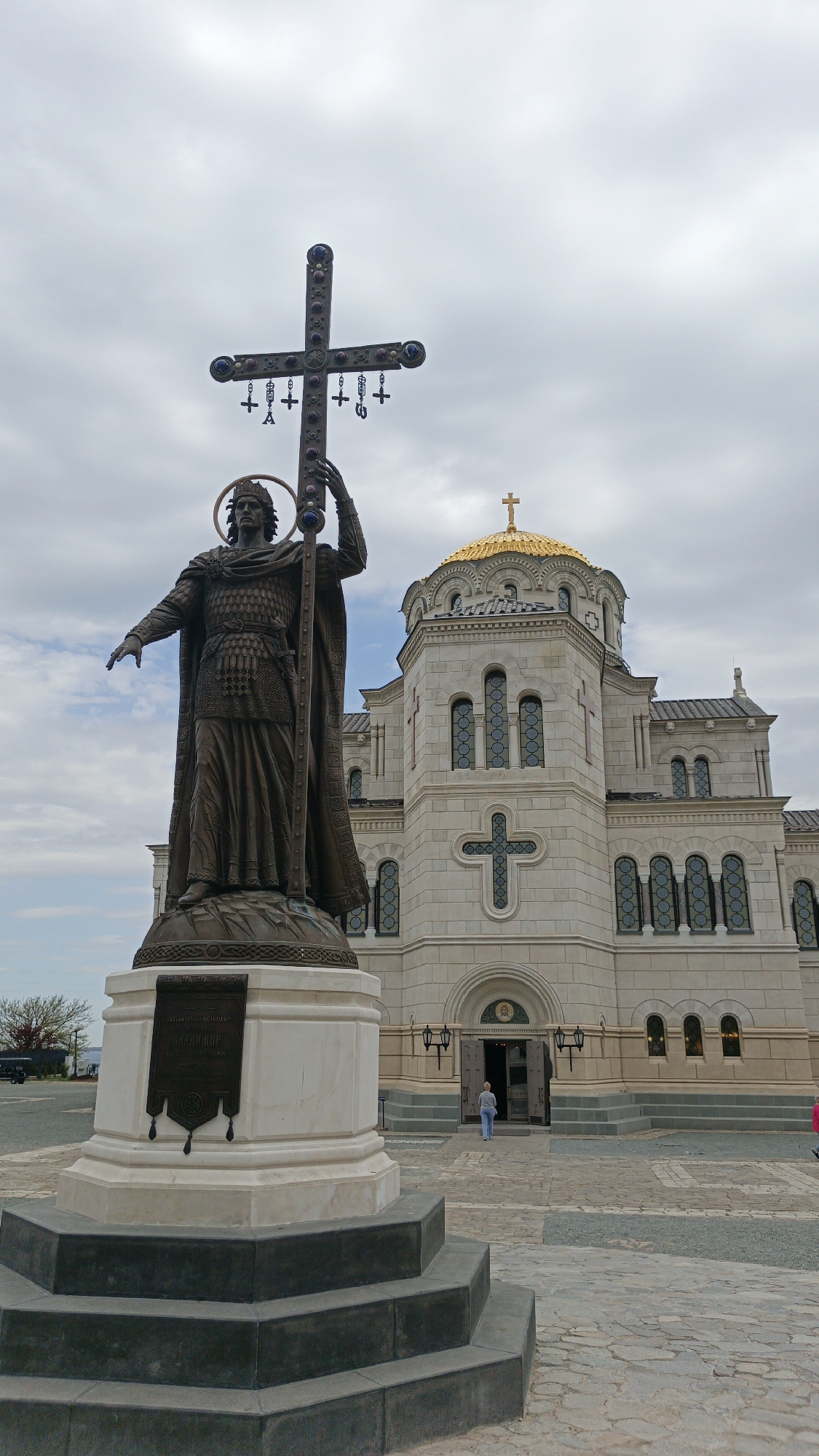 A statue of a man holding a large cross stands in front of a building with a golden dome in Sevastopol, Ukraine.
The statue is made of bronze and depicts a man with a halo around his head. He is wearing a long robe and armor, and he is holding a large cross in his right hand. The cross has several smaller crosses hanging from it. The statue is standing on a white marble base. There is a plaque attached to the front of the base.
The building in the background is a large, white stone church with a golden dome. The church has many windows and crosses carved into its exterior walls.
In front of the church, a woman is walking towards the entrance.
The sky is overcast with gray clouds.