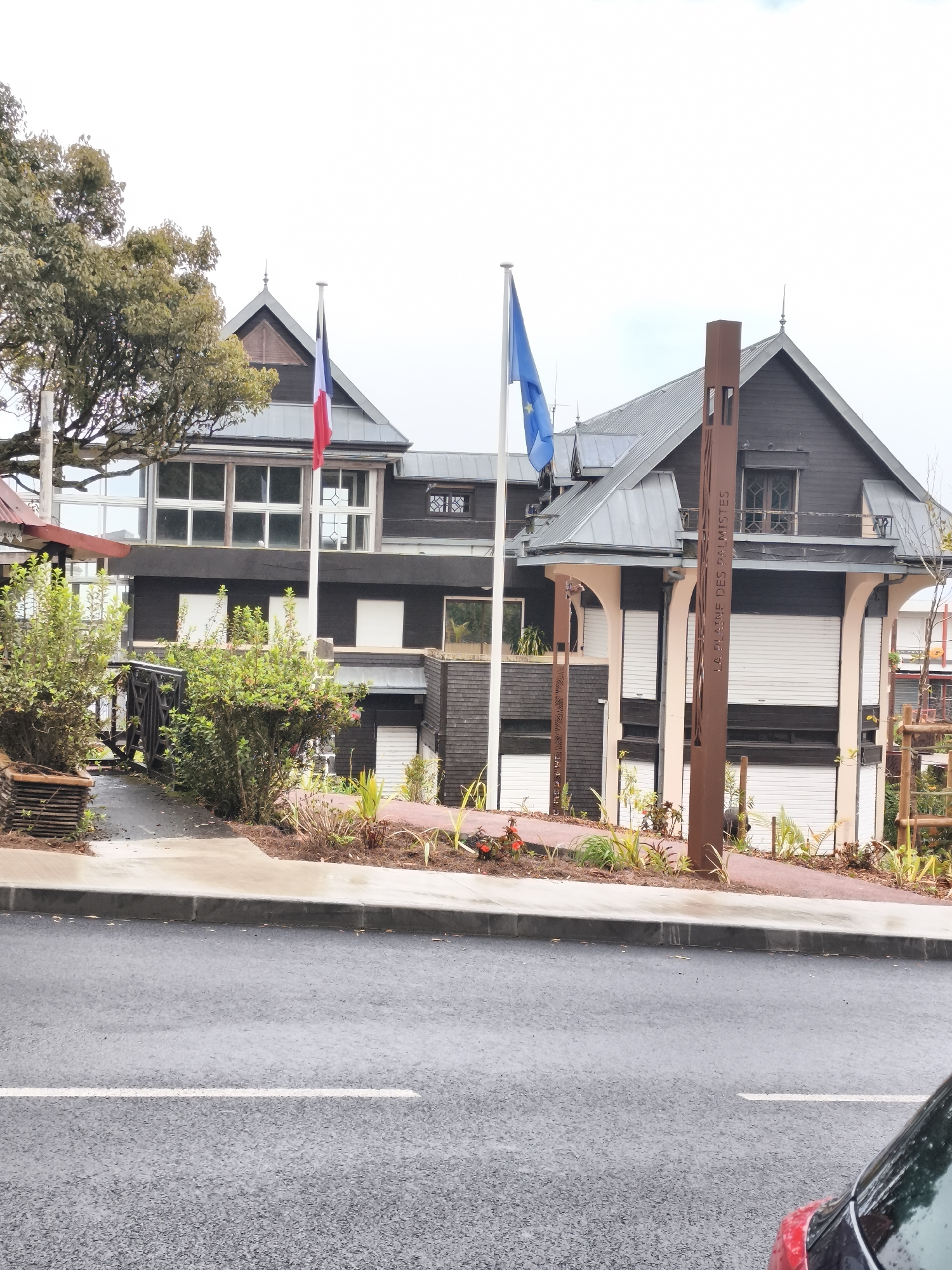 The image depicts the exterior of a multi-story building in La Plaine-des-Palmistes, France. The building is dark wood with a gray metal roof and features numerous large windows. Two flags, one French and one European, are flown from poles in front of the building. A large, stylized metal post with text on it stands in the foreground. The area in front of the building is landscaped with plants and a paved walkway. A road with a white line marking is visible in the lower part of the image, and the rear of a red car is visible in the bottom right corner. The sky is overcast.