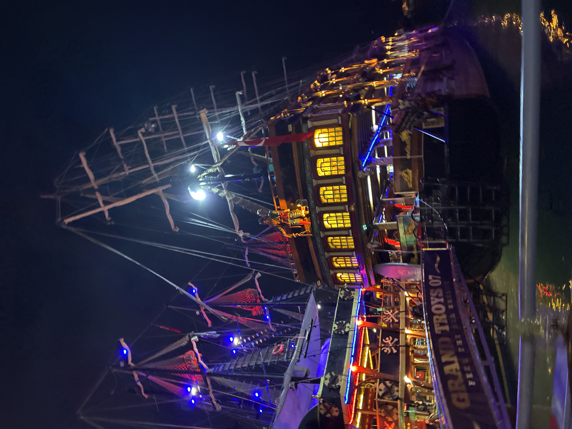 The image depicts a brightly illuminated, ornate pirate-themed ship docked at night, likely in Alanya, Turkey, given the location context. The ship is heavily decorated with colorful lights, including blue, red, and yellow, accentuating its intricate details.
The ship features multiple decks, masts, and rigging, giving it a grand and imposing presence. Windows on the ship's structure glow with a warm yellow light, suggesting an interior that is either occupied or illuminated. Flags and banners are visible, adding to the festive and somewhat theatrical atmosphere. The water around the ship reflects the lights, creating a shimmering effect.
On the side of the ship, a prominent banner displays text, partially obscured but clearly readable as "GRAND TROYS 07". Below this, more text is visible, though less distinct. The overall impression is one of a lively evening at a harbor, possibly a tourist attraction or a venue for evening entertainment. The night setting and the abundance of lights create a magical and inviting ambiance. The scene appears calm, with the ship stationary at its dock.