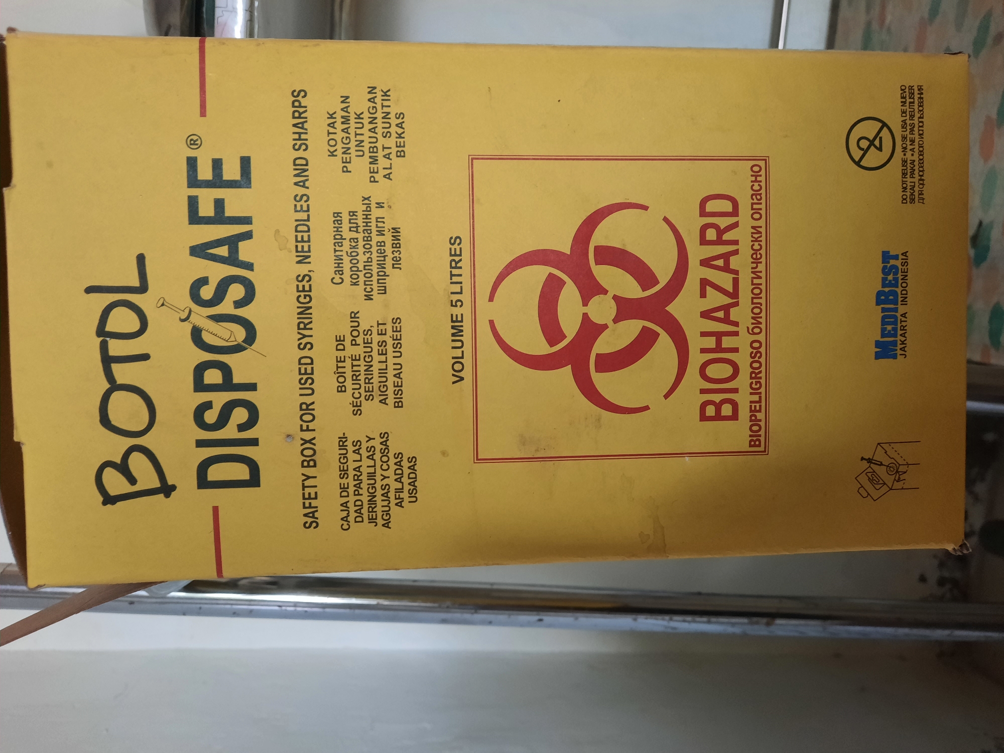 The image shows the side of a yellow disposal box labeled "BOTOL DISAPOSAFE" in large black letters. The box is designed for used syringes, needles, and sharps. It features multiple language translations for its purpose, including Spanish, French, Russian, and Malay. Prominently displayed is the biohazard symbol in red, with the word "BIOHAZARD" below it, followed by "BIOPELIGROSO биологически опасно". The box also indicates a volume of 5 liters. The brand name "MediBEST JAKARTA INDONESIA" is visible at the bottom, along with a recycling symbol indicating "DO NOT REUSE". The box appears to be in a clean, indoor environment, possibly a medical facility or waste disposal area. There are no people visible in the image. This disposal box is not specific to Lhokseumawe, Indonesia, as the brand "MediBEST" is associated with Jakarta, Indonesia.