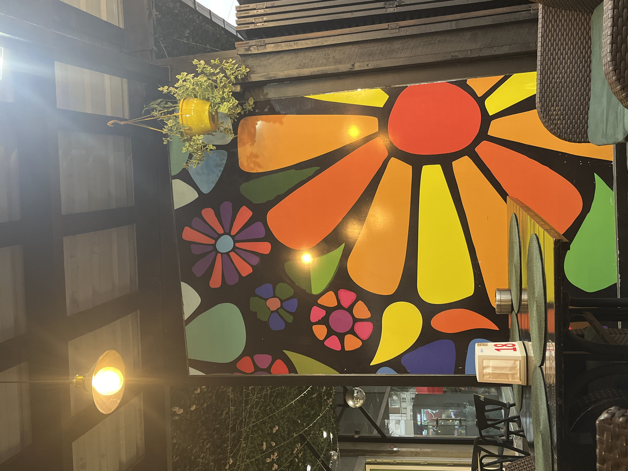 The image shows a brightly decorated outdoor seating area, likely a cafe or restaurant, in Kobuleti, Georgia. Dominating the background is a large, vibrant mural of a stylized flower with bold, colorful petals set against a black background. The flower design incorporates shades of orange, yellow, red, and green. Above the mural, a pendant light fixture with a warm yellow glow hangs from the ceiling. A yellow potted plant with trailing greenery is suspended nearby.
In the foreground, a black table is set with green placemats. On the table, a white napkin dispenser displays the number "18" in red numerals, and a metal ashtray sits on another placemat. Dark wicker chairs are visible around the table, and a portion of a wicker armchair with a green cushion is in the lower right corner. In the background, through a glass storefront, other tables and chairs can be seen, along with glimpses of the street. The overall atmosphere suggests a casual and lively dining or gathering space.