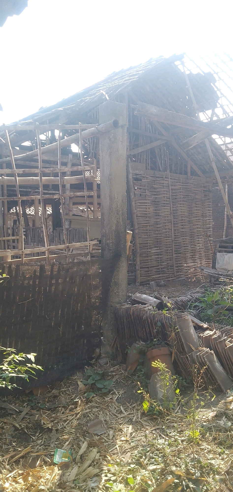 The image depicts the exterior of a dilapidated building, likely a traditional Indonesian structure, possibly located in Plemahan, Indonesia. The building is characterized by its exposed bamboo framework and woven bamboo walls, with a partially collapsed thatched roof. Several beams and supports are visible, some appearing to be in disrepair. In the foreground, a pile of clay roof tiles is stacked on the ground, alongside scattered debris consisting of dried leaves, twigs, and what appears to be broken pieces of bamboo or wood. Some greenery, including small plants and vines, is growing around the base of the structure and among the debris. The overall scene suggests an abandoned or under-construction site, exposed to natural elements under a bright, overcast sky. There are no people visible in the image.