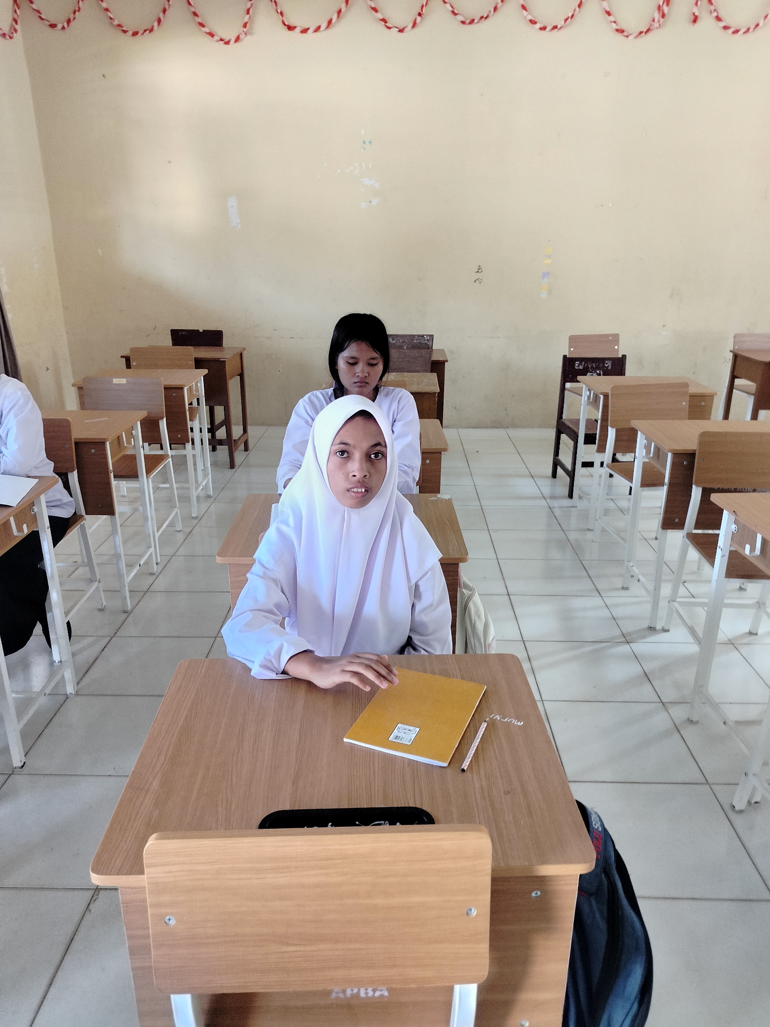 The image depicts two young females, likely students, seated at wooden desks in a classroom. The student in the foreground, wearing a white hijab and a white long-sleeved top, is looking directly at the camera with a neutral expression. Her hands are resting on the desk in front of her, on which a yellow notebook and a pen are placed. To her left, another student, also wearing a white long-sleeved top, is visible from the chest up, looking down. The classroom environment appears simple, with rows of similar desks and chairs. The walls are painted a light, neutral color, and a festive red and white garland is strung along the top of the wall. The floor is tiled. The overall impression is of an ordinary school day in what is likely a public school in Lawebekung, Indonesia, given the context of Indonesian culture and school settings.
