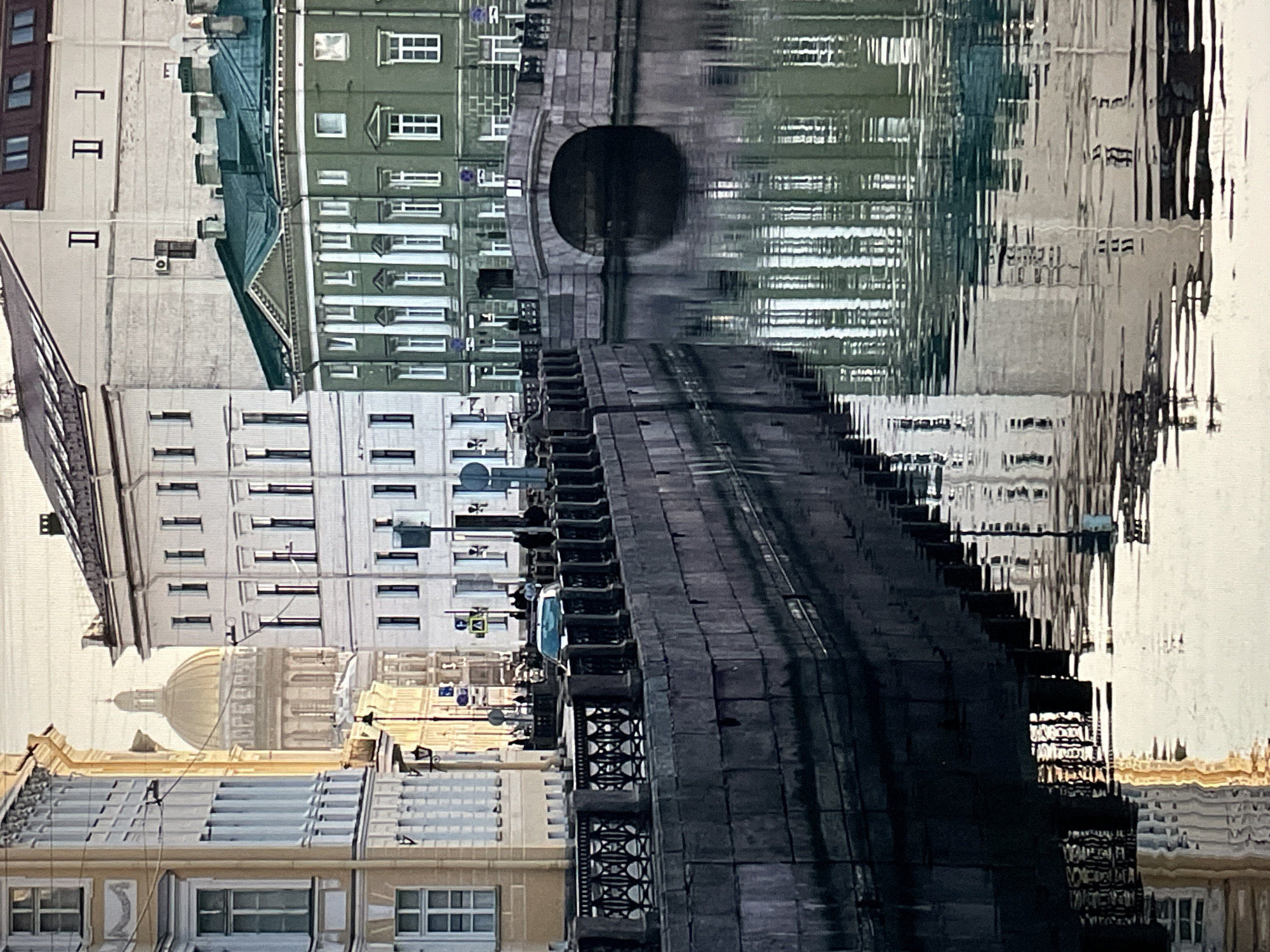 This image depicts a tranquil urban scene along a canal in Saint Petersburg, Russia. The most prominent distant landmark is the golden dome and upper rotunda of St. Isaac's Cathedral, visible in the background, centered between buildings.
In the foreground, the calm water of a canal occupies the lower half of the frame, displaying clear, distorted reflections of the surrounding architecture and sky. The left bank of the canal features a dark grey, rough-hewn stone embankment, topped with a black wrought-iron railing composed of decorative scrollwork panels between solid posts. Behind this railing, a street or pavement is visible, where several indistinct figures (pedestrians) are walking, appearing as dark silhouettes. A dark-colored vehicle is also present on this street, partially obscured by the railing. To the left, a multi-story, light yellow classical building with white window frames and ornate details lines the canal.
In the midground, an arched stone bridge spans the canal, connecting the left bank to the right. Beyond the bridge on the right bank stands a distinctive multi-story green classical building featuring white pilasters, a pediment, and a dark green roof. Other multi-story buildings, varying in color from white to cream, line the canal banks further back. Streetlights and various traffic signs are visible along the streets.
The scene is illuminated by soft, directional light, suggesting either early morning or late afternoon, which highlights the golden dome and the facades of the buildings while creating gentle reflections on the water. No direct interactions between individuals are discernible.