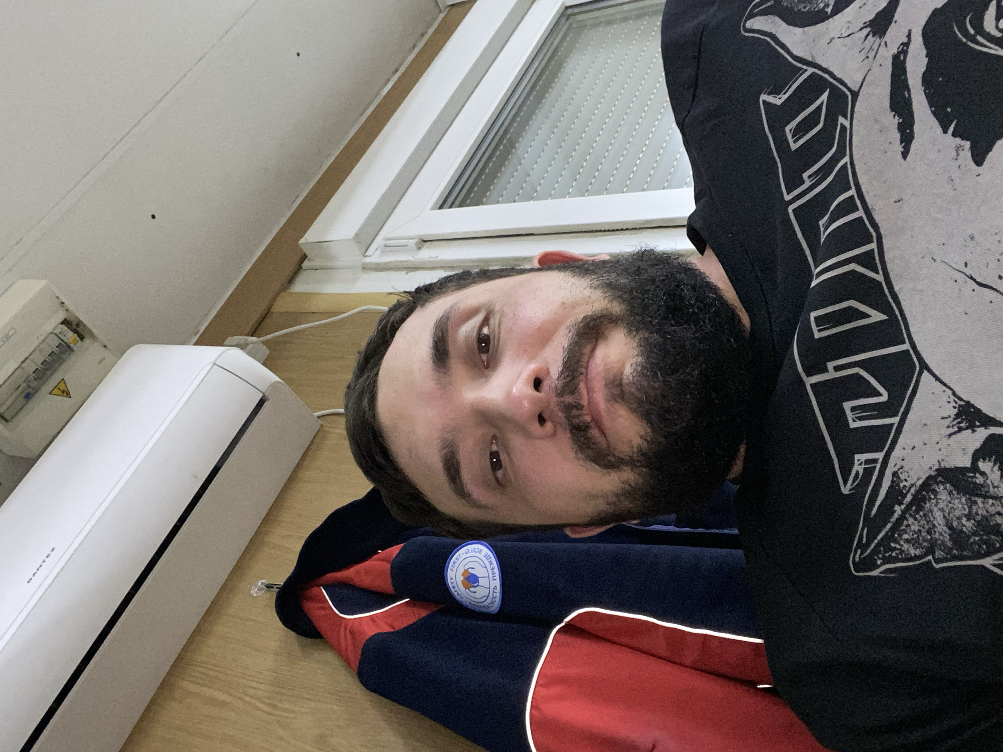 A man with a beard and dark hair is taking a selfie indoors. He is wearing a black t-shirt with a graphic print. Behind him, a navy blue and red jacket is hanging on a wall. Above the jacket, a white air conditioner is mounted on the wall. To the right of the man, a white-framed window with blinds is visible. The man is positioned in the foreground, with the background elements slightly out of focus. The overall impression is a casual, personal snapshot.