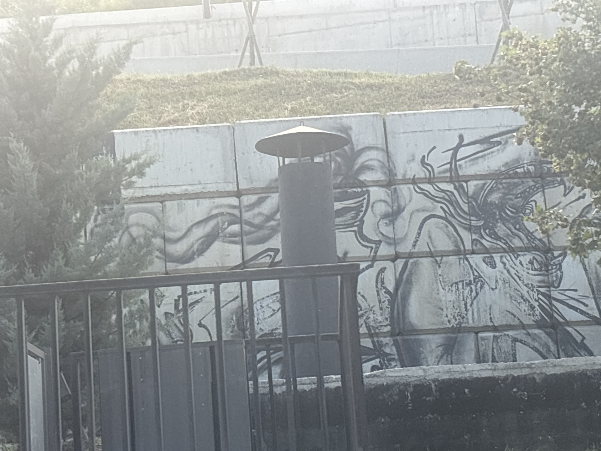 The image shows a grey metal chimney with a conical cap, positioned in front of a concrete wall adorned with a black and white mural. The mural depicts stylized figures with flowing lines, resembling mythical creatures or abstract human forms. To the left of the chimney, a green evergreen tree is visible, and to the right, a tree with green foliage. In the foreground, a black metal fence with vertical bars runs across the bottom of the frame. The background consists of a grassy slope and further concrete structures, suggesting an outdoor urban environment. The lighting is hazy, potentially indicating early morning or late afternoon. The scene is located in Tbilisi, Georgia, specifically at the St. Andrew the First-Called Georgian University of the Patriarchate of All Georgia, which features a prominent mural on its exterior walls.