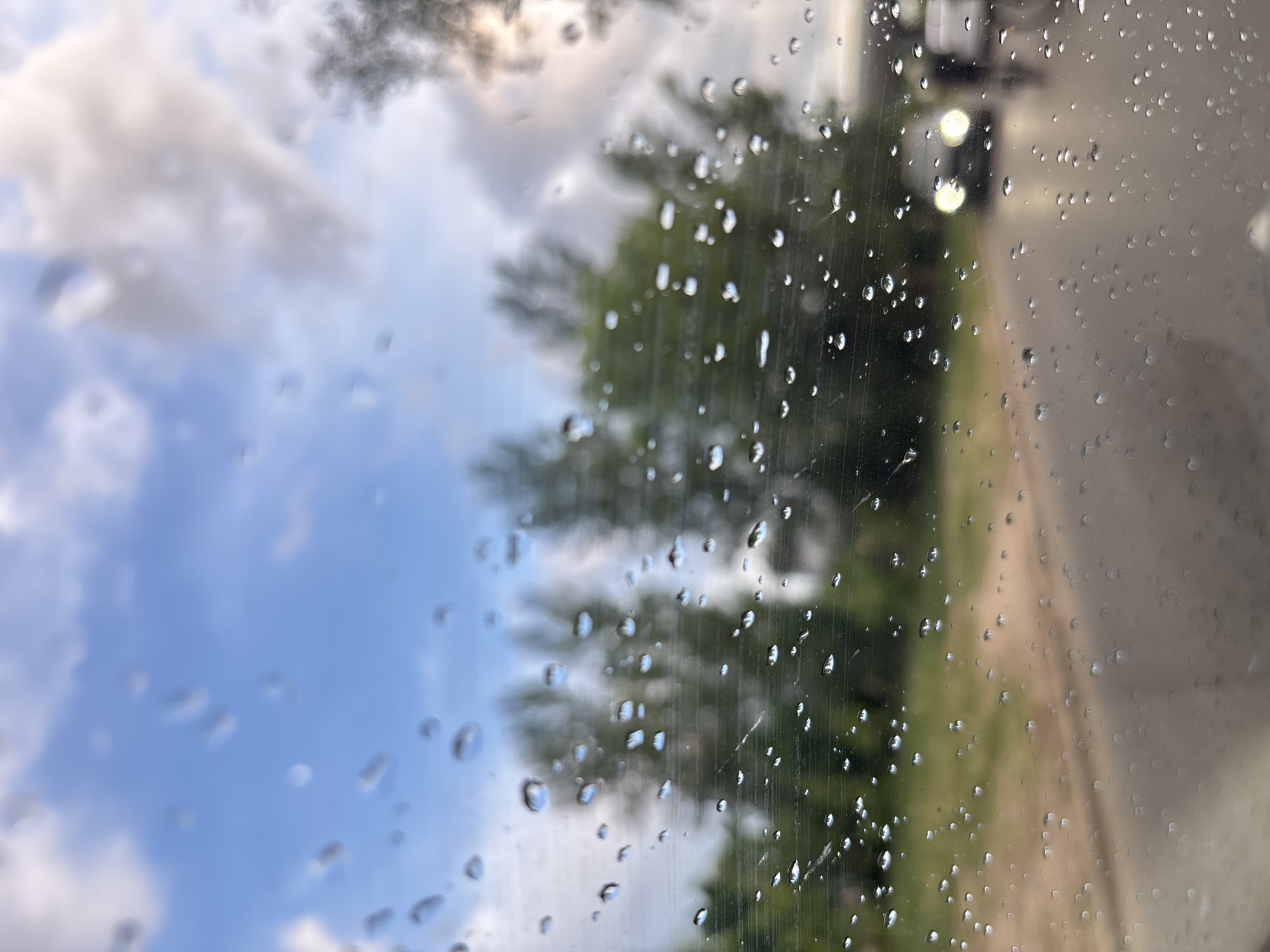 The image shows a view through a rain-splattered window. Outside, the sky is partly cloudy with patches of blue sky visible. A blurred outline of trees can be seen in the middle ground. To the right of the trees, a street is visible, with a blurred vehicle or object in the background, possibly on the roadside. The location is unidentified.